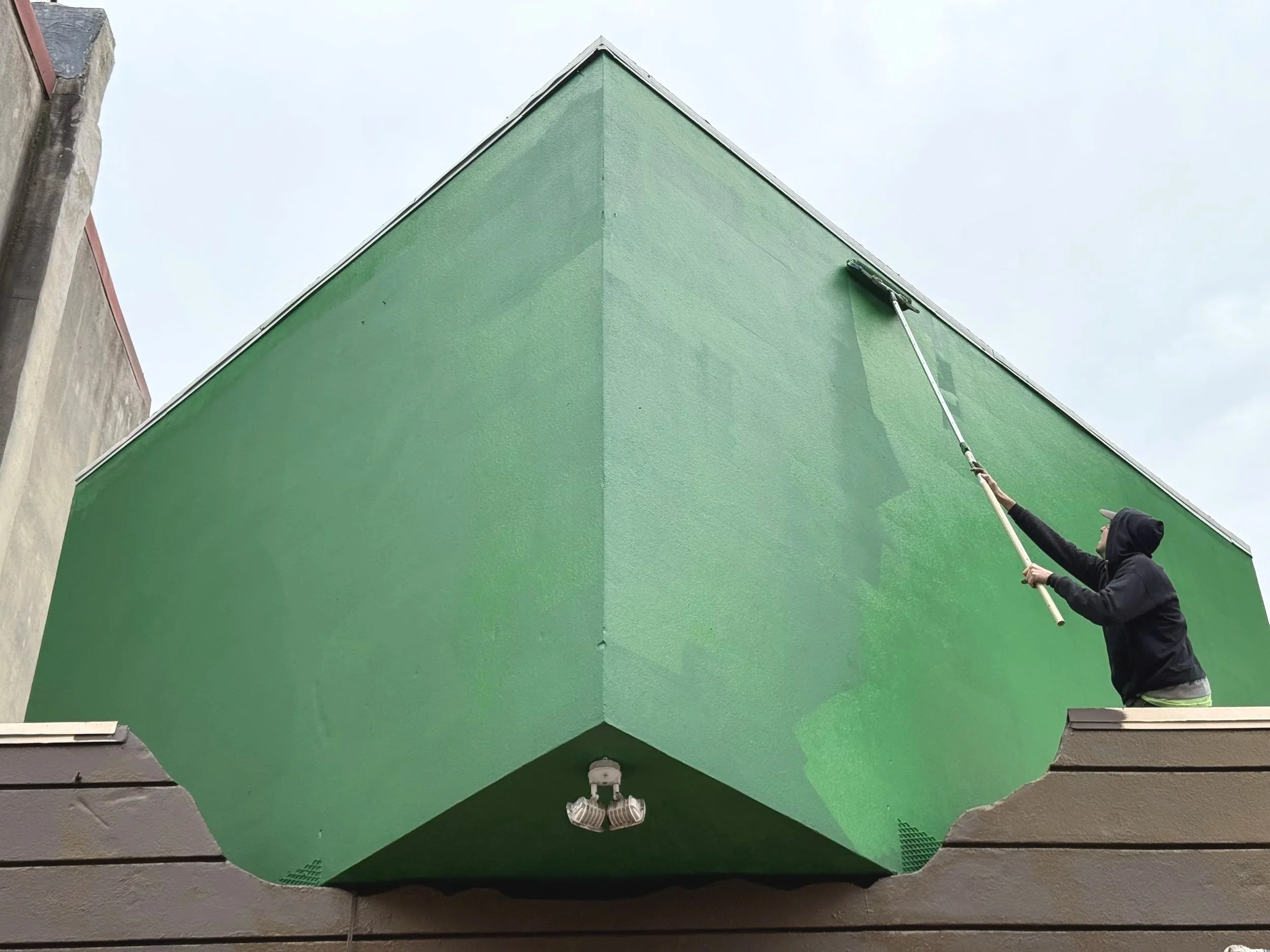 Person in a hoodie cleaning the exterior wall of a small green building with a squeegee on a pole.