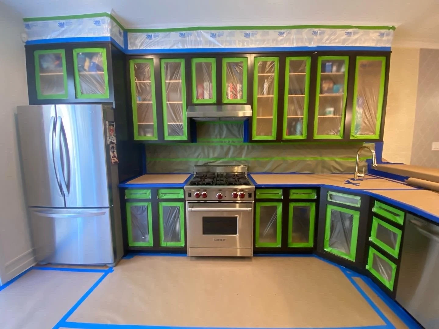 Kitchen cabinets with blue tape and plastic covers, stainless steel refrigerator, gas stove, and sink.