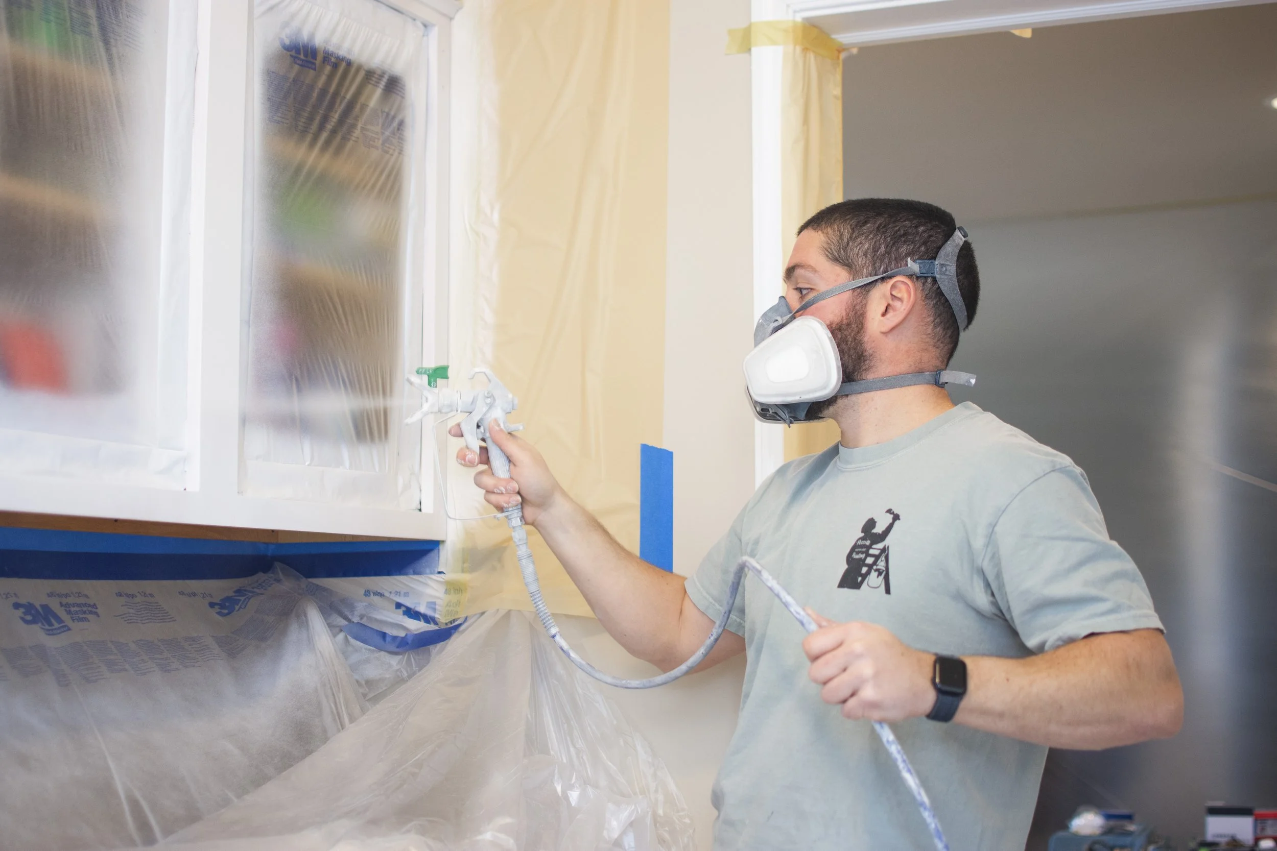 A man wearing a respirator mask and gray t-shirt spray painting a cabinet in a room with plastic protective covering.