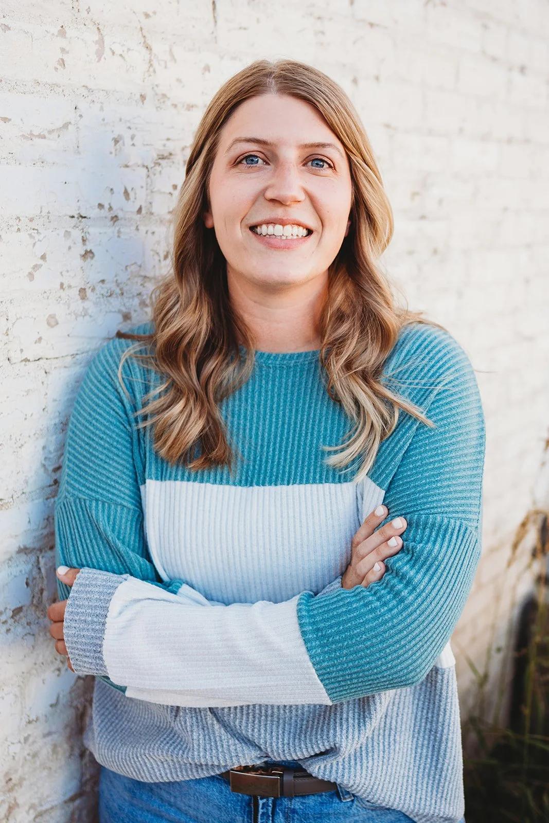 A young woman with long wavy hair and blue eyes smiling, standing against a white brick wall, wearing a blue and white striped sweater.