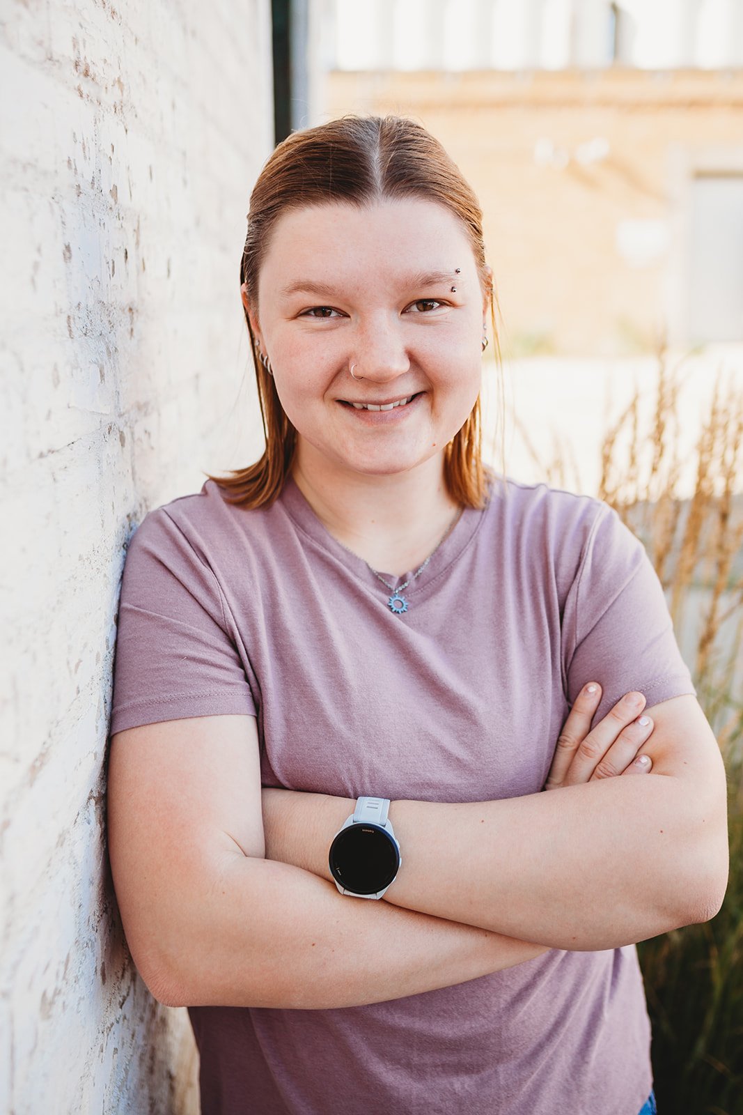 A young woman with red hair leaning against a white brick wall, smiling, wearing a pink T-shirt, a necklace, earrings, a nose ring, an eyebrow piercing, and a smartwatch.