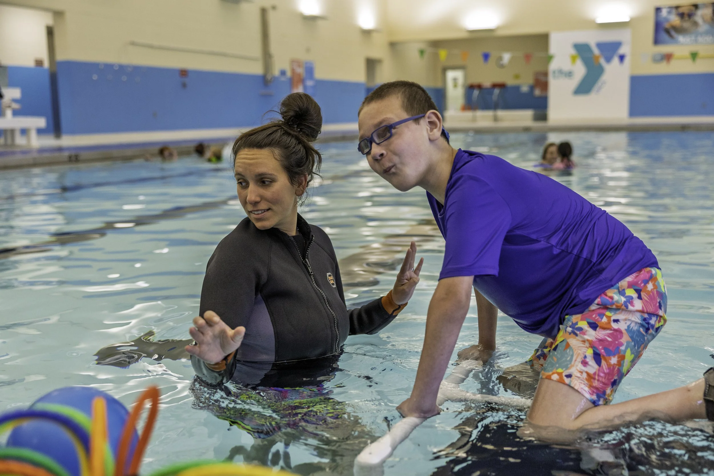 patient in colorful swim trunks and goggles speaks with a therapist in the pool, with other swimmers in the background at an indoor aquatic center.