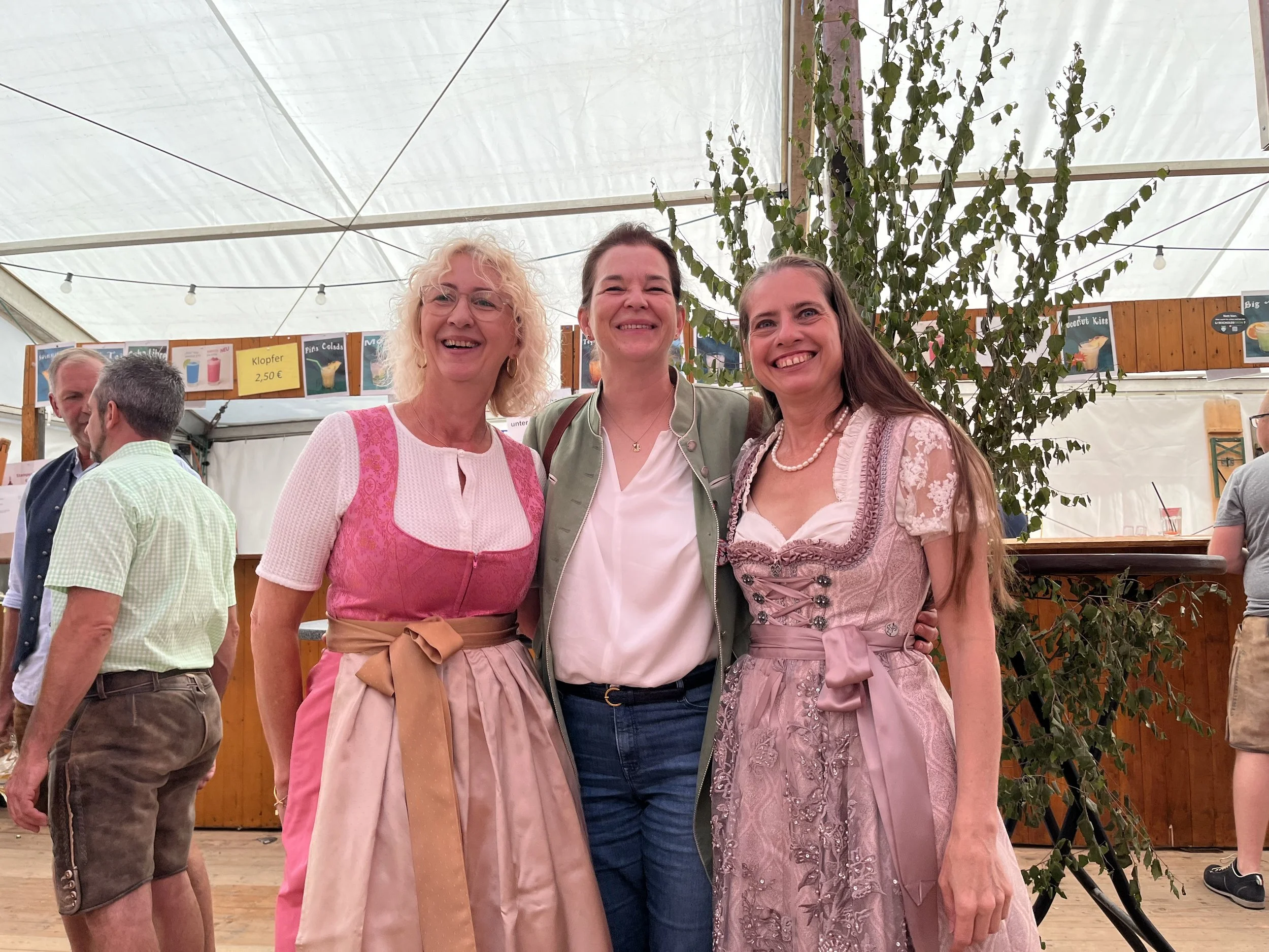 Three women in traditional dirndls and a woman in modern clothes smile and pose together inside a tent at a festival, with other people and a wooden bar in the background.