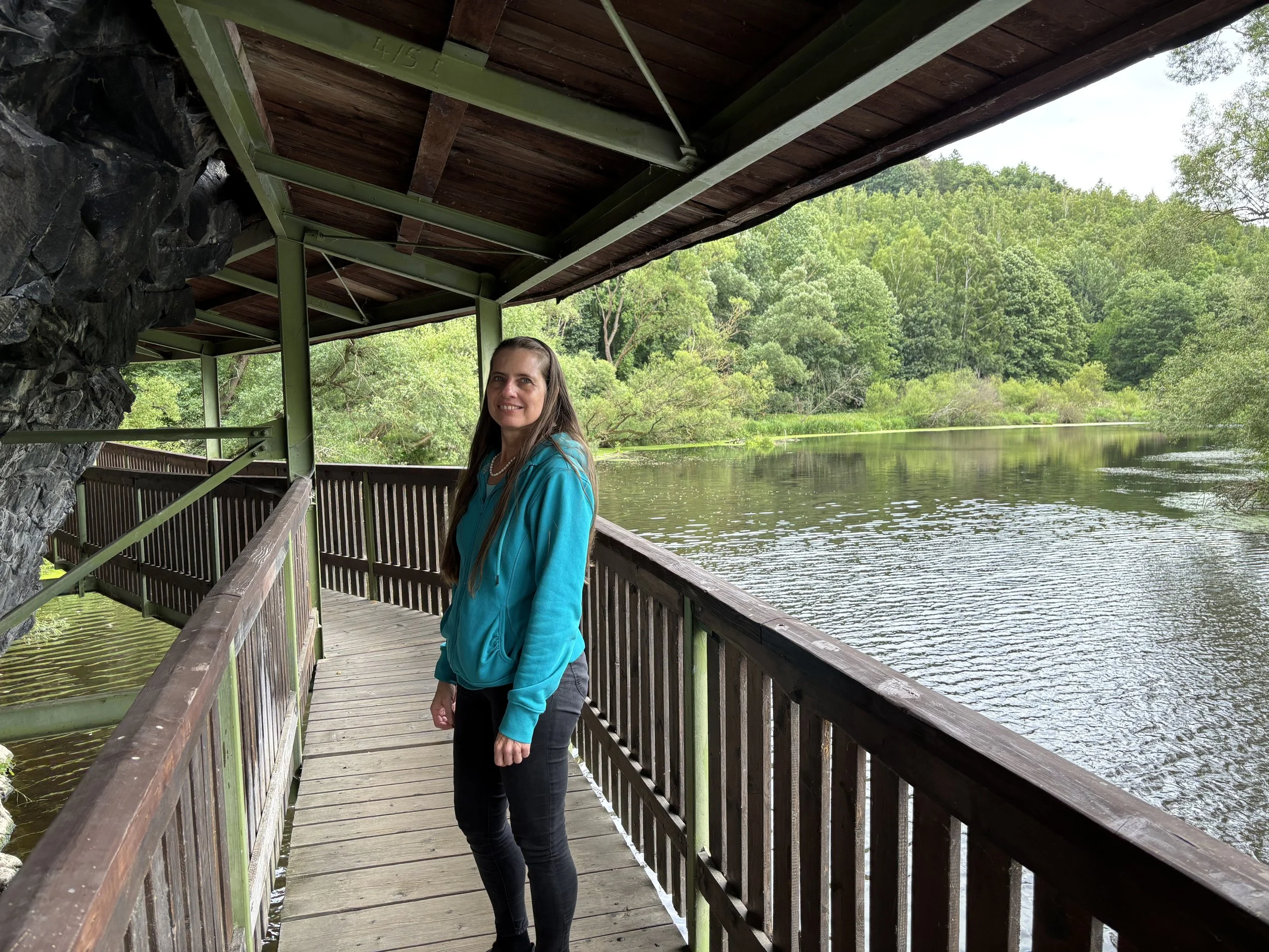 Katrina in a blue jacket standing on a wooden walkway next to a river, surrounded by green trees.