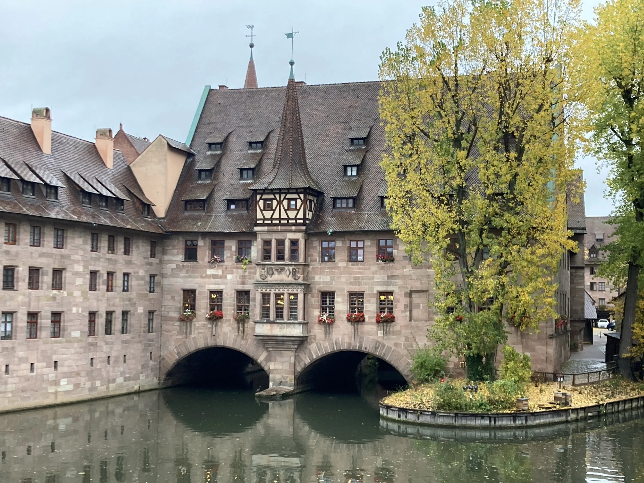 Old European building with timber framing and a steep tiled roof, situated by a waterway with a small island and trees with yellow leaves in the foreground.