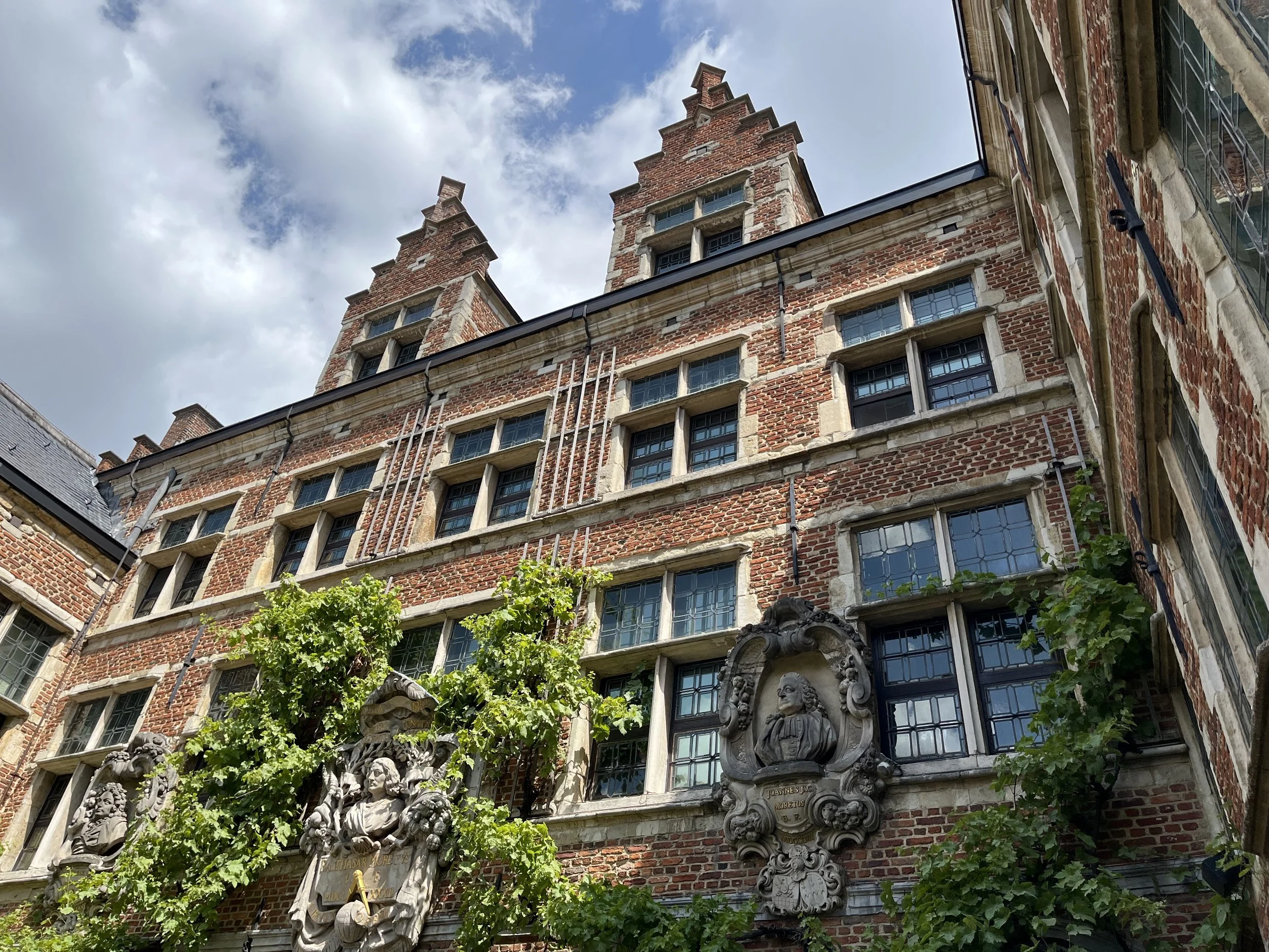 A historic brick building with ornate statues and greenery in the foreground, under a partly cloudy sky.
