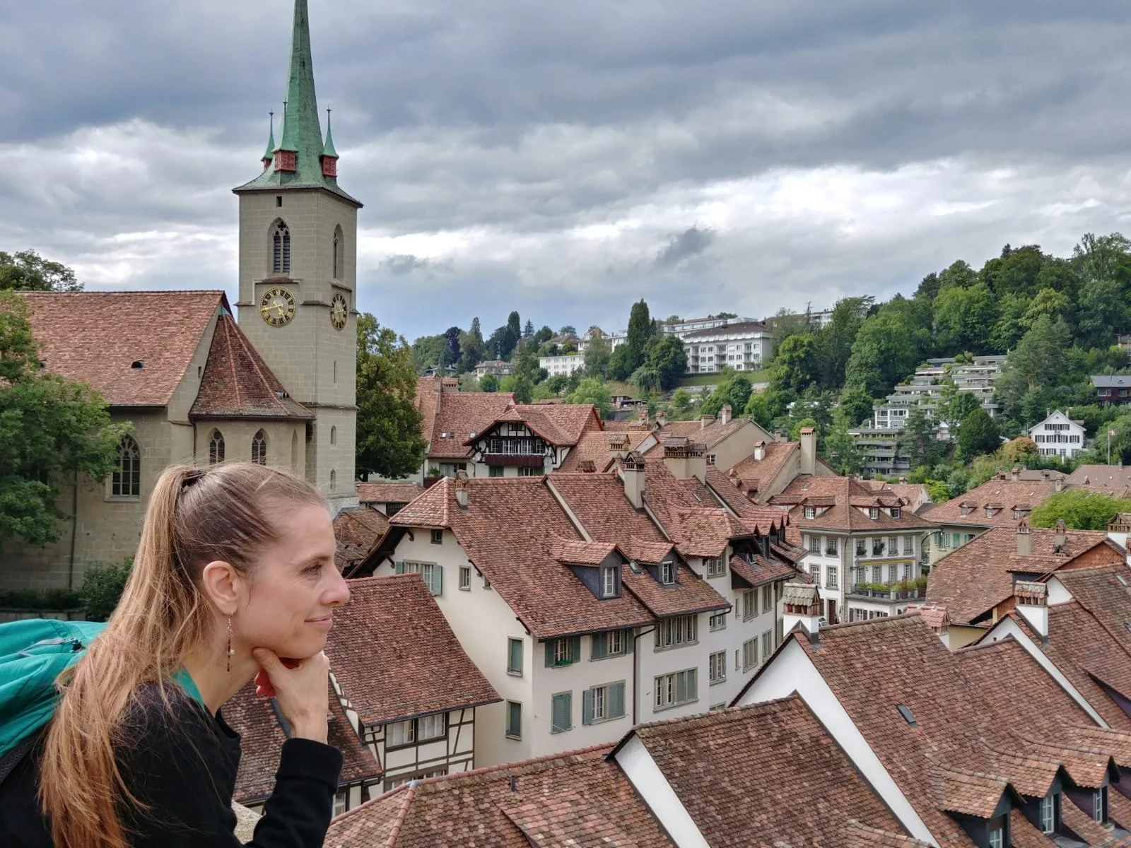 Katrina, wearing a black jacket and backpack, rests her chin on her hand while looking over a European town with red-tiled roofs, a church with a tall spire, and lush green hills under a cloudy sky.