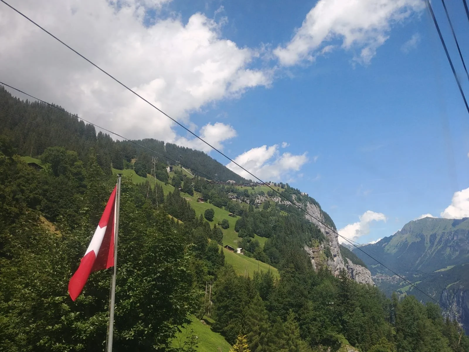Scenic view of a Swiss landscape with lush green hills, rocky cliffs, and a bright blue sky with some white clouds. A Swiss flag is visible in the foreground, and power lines cross the sky.