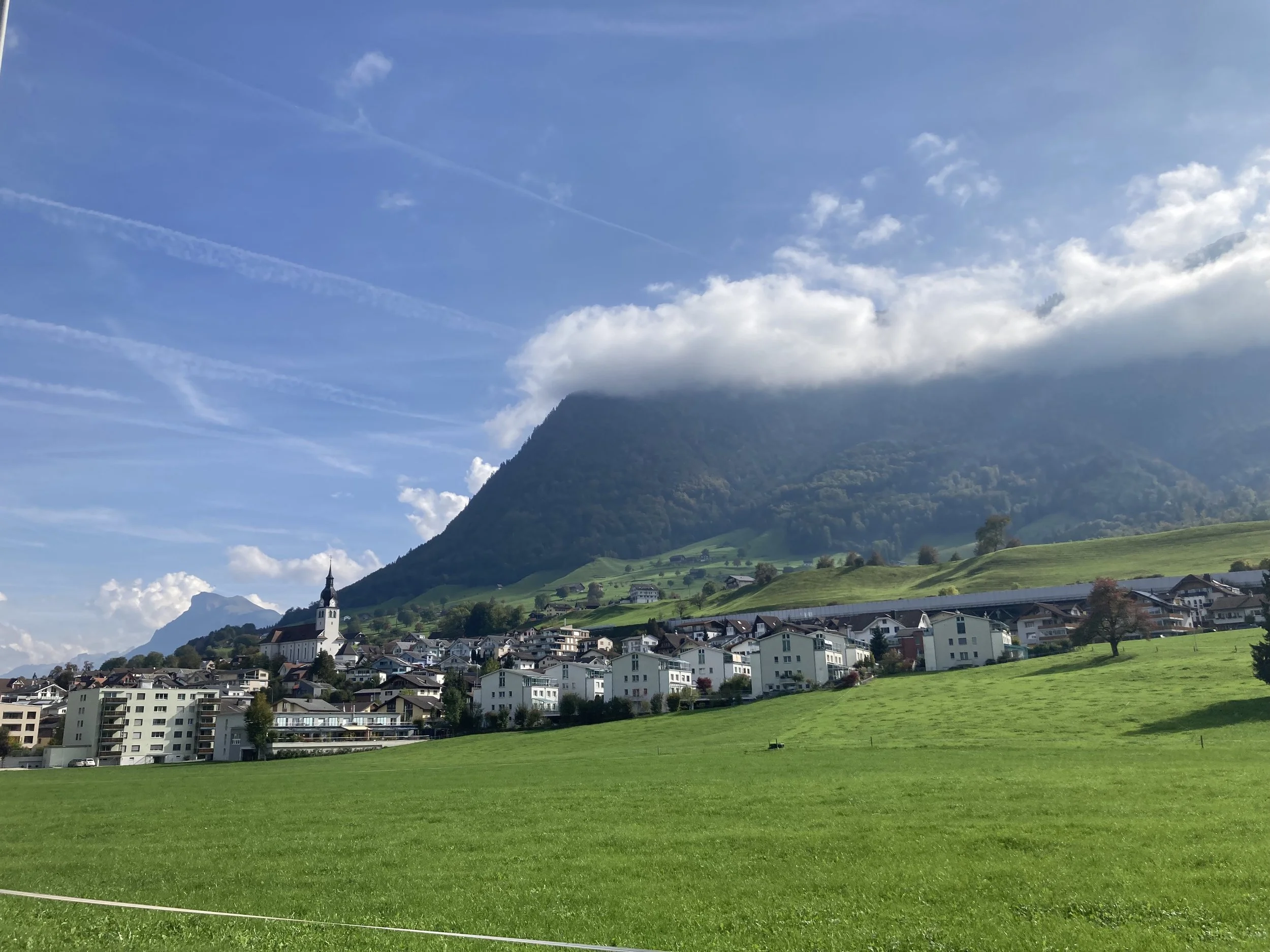 A scenic view of a small town in a valley with green fields, residential buildings, and a church with a tall steeple. Surrounding mountains are partly covered with clouds under a partly cloudy sky.