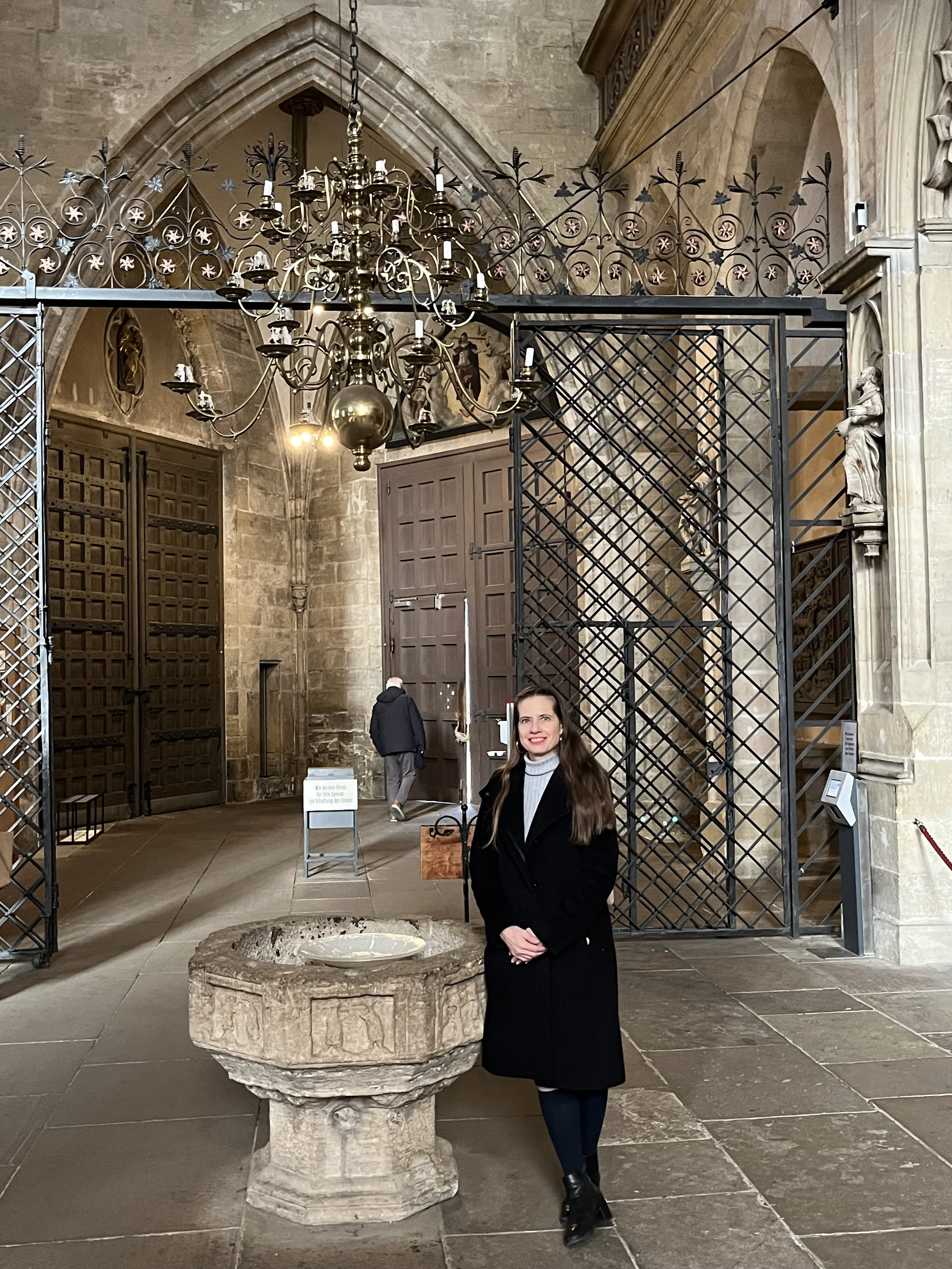 A woman with long brown hair smiling, dressed in a black coat, standing inside a historic church or cathedral, with a stone baptismal font in front of her, a grand chandelier hanging above, and architectural details like statues and arches around.