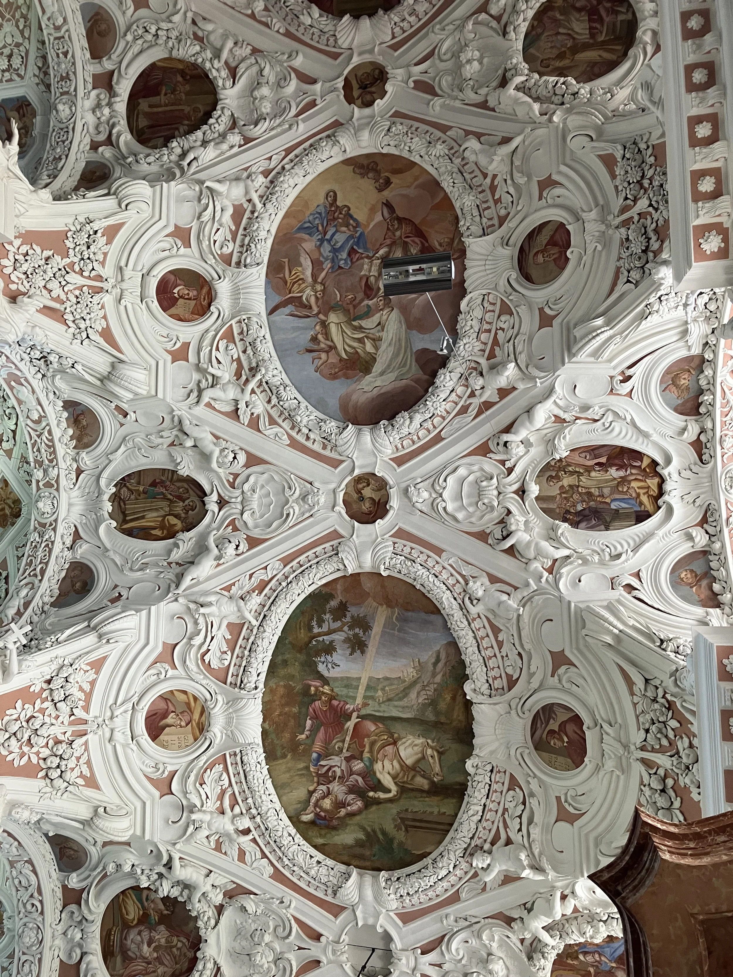 Ceiling frescoes with religious scenes and elaborate white stucco decorations in a church ceiling.