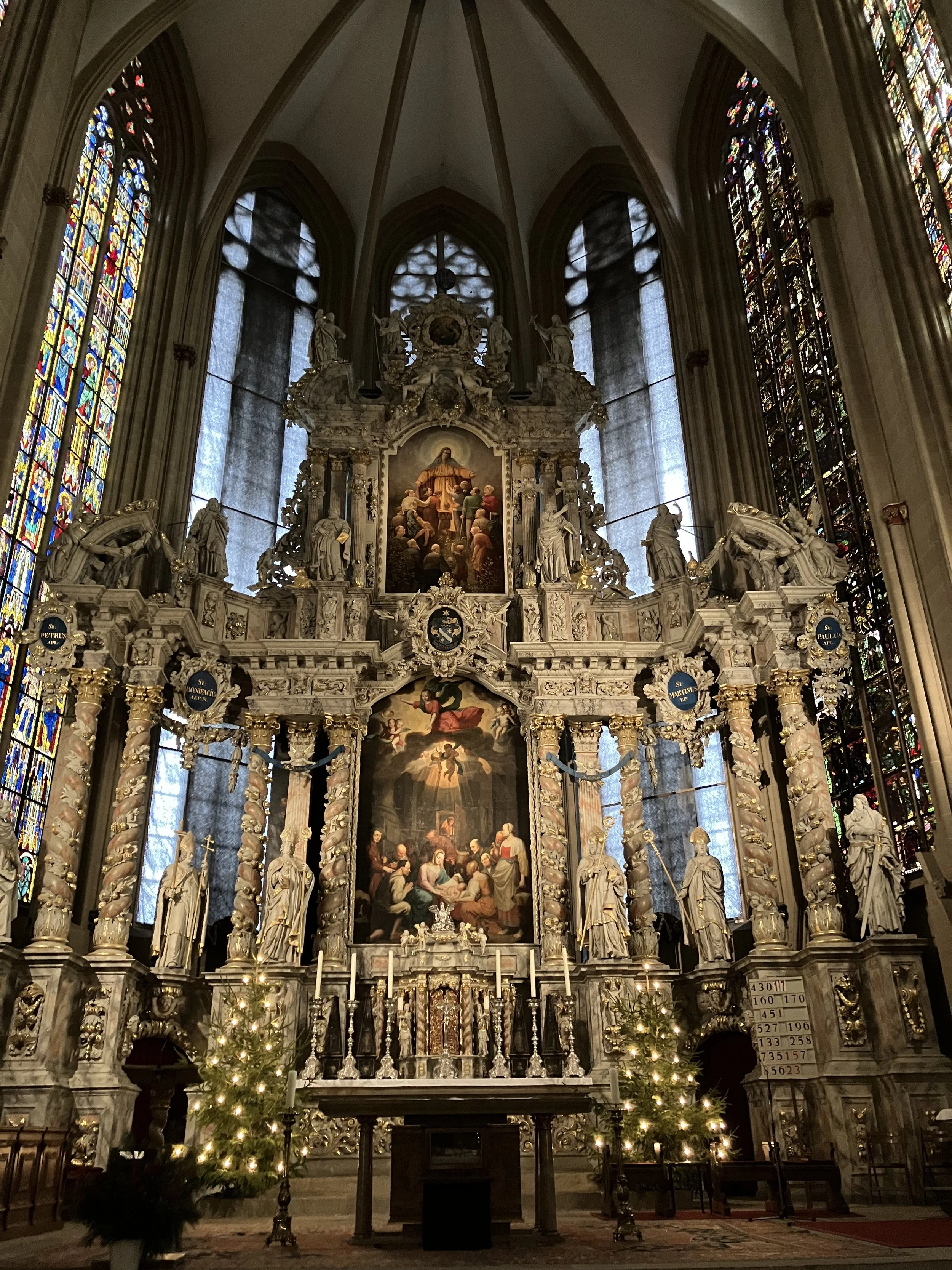 Interior of a church with ornate altar, statues, large religious paintings, stained glass windows, and Christmas trees with lights.