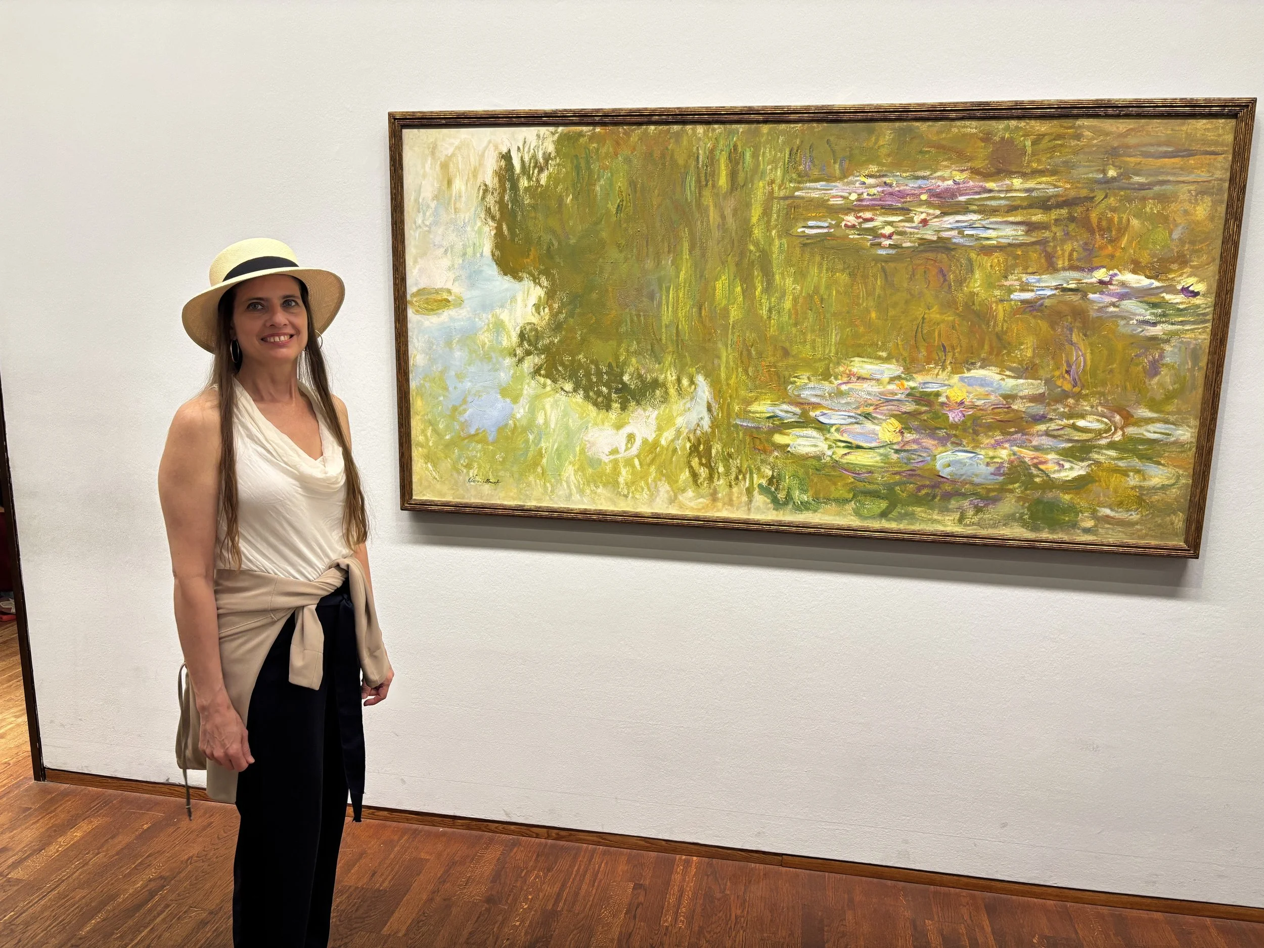 Katrina wearing a beige sunhat, white sleeveless top, and black trousers standing next to a large Impressionist painting of a water lily pond in an art gallery.