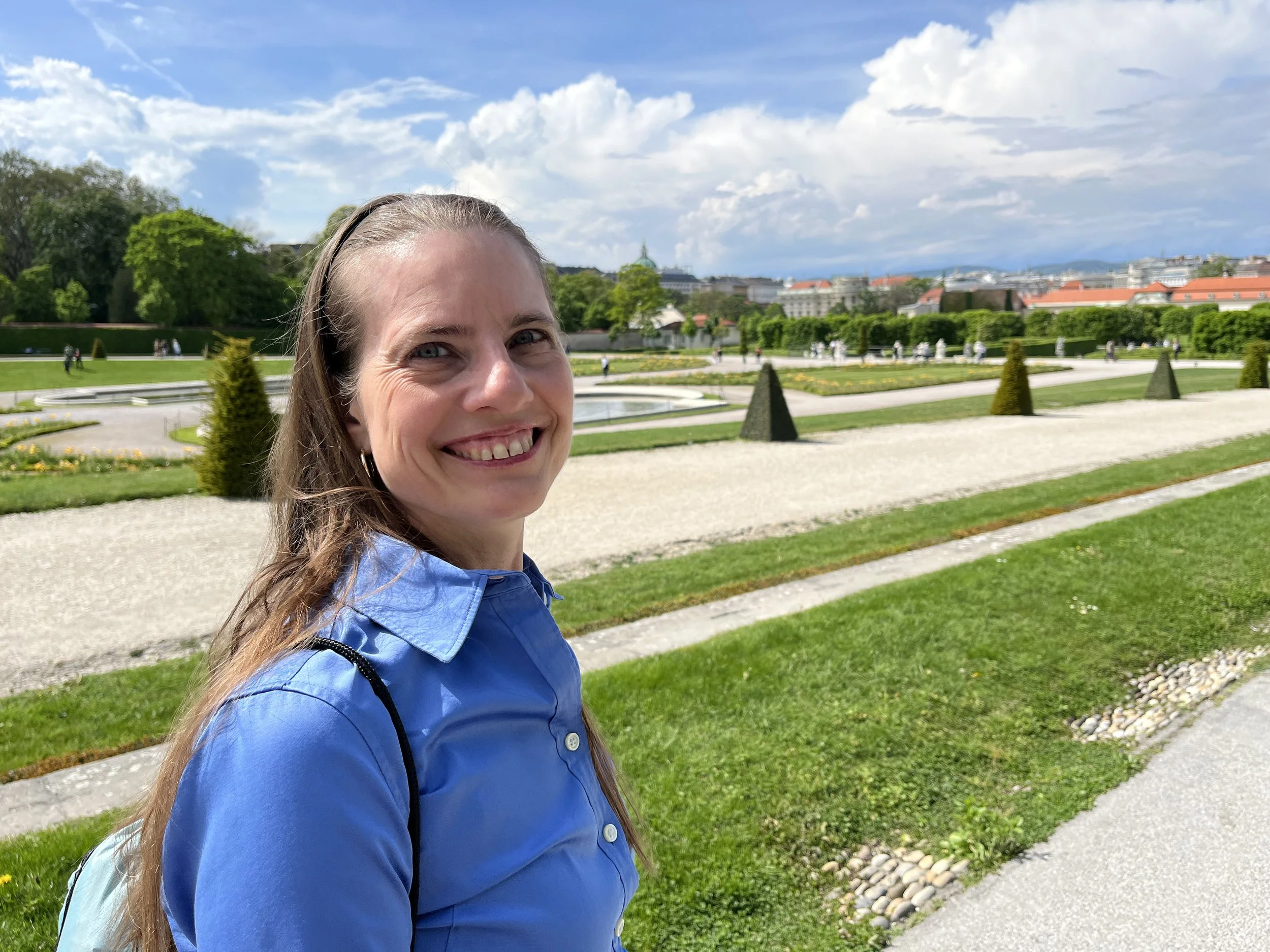 Katrina smiling outdoors in a park with neatly maintained garden beds, gravel paths, manicured lawns, and historic buildings in the background under a partly cloudy sky.
