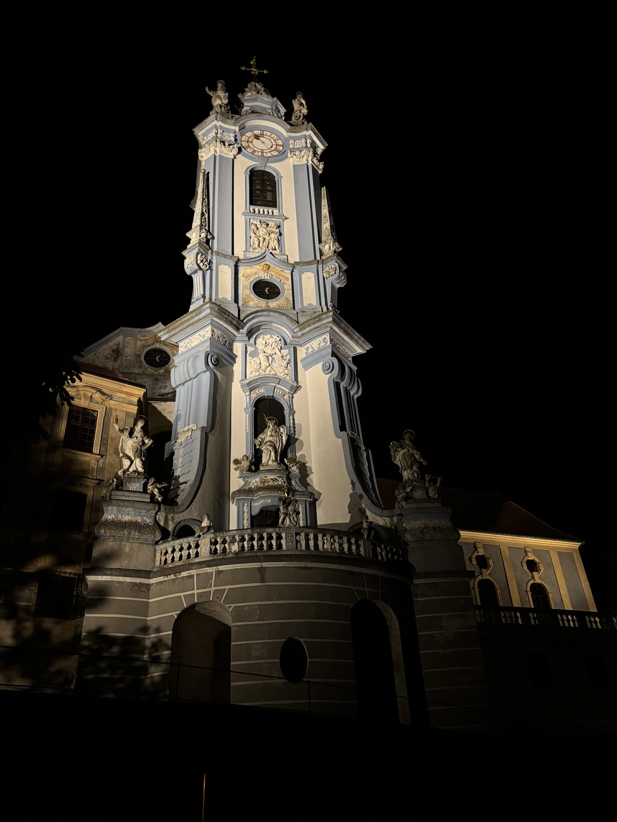 Night view of a historic church with baroque-style architecture, featuring ornate sculptures, a clock tower, and decorative details illuminated against a dark sky.
