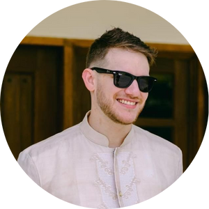 Young man with short hair and a beard wearing sunglasses and a white traditional shirt, smiling indoors.