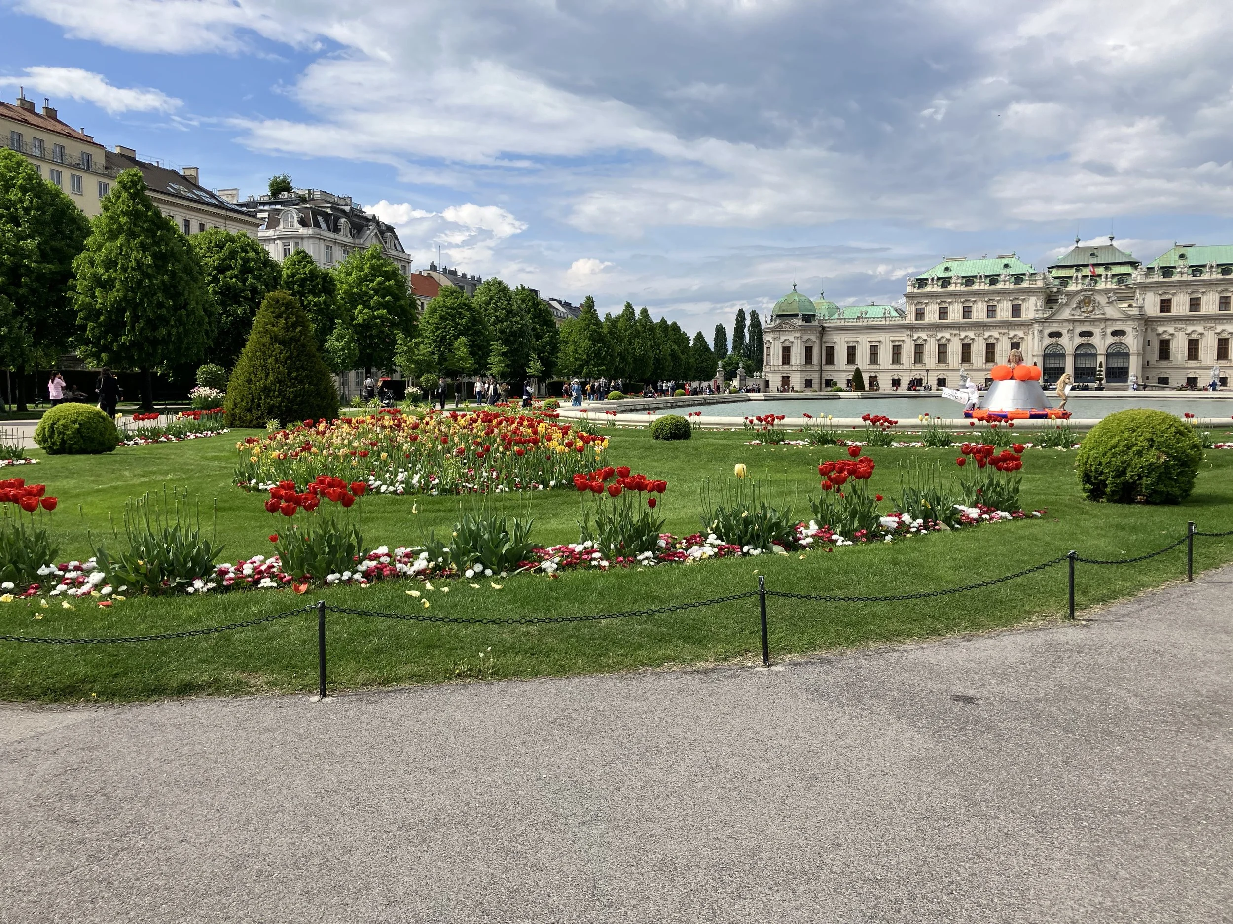 A city park with a flower garden featuring red, yellow, and white tulips, surrounded by green lawn and trees, with a historic building in the background under a partly cloudy sky.