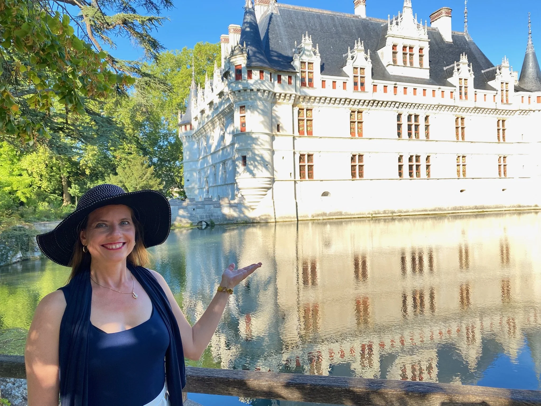 Katrina with a large black sun hat, blue top, and gold jewelry standing in front of a castle by a moat, smiling and gesturing toward the castle.