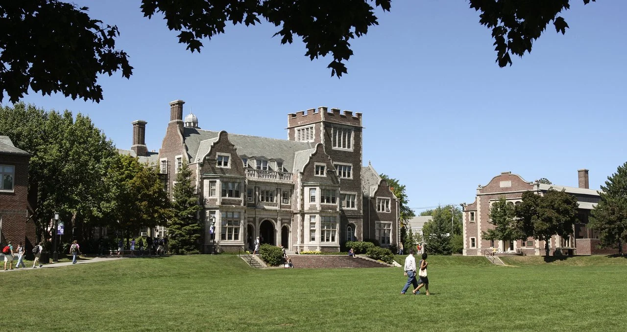A historic castle-like building with multiple turrets and decorative brickwork, surrounded by trees and a well-maintained lawn, with people walking on the paved pathways.