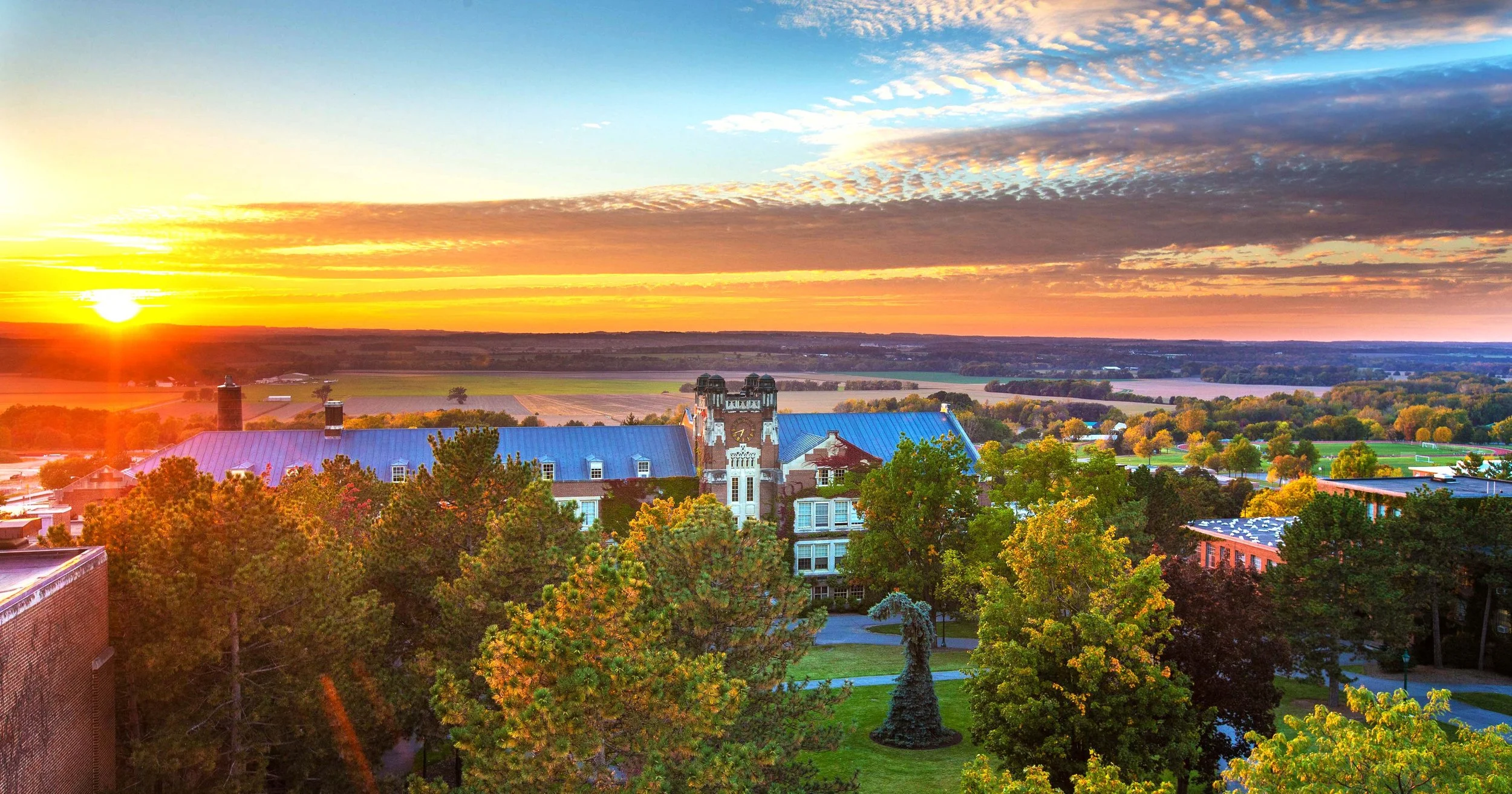 Sunset over a campus with trees and historic buildings, a blue-roofed structure and a tower.