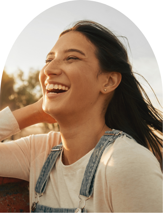 A woman smiling and enjoying outdoors with her hand behind her head, wearing a white shirt and denim overalls.