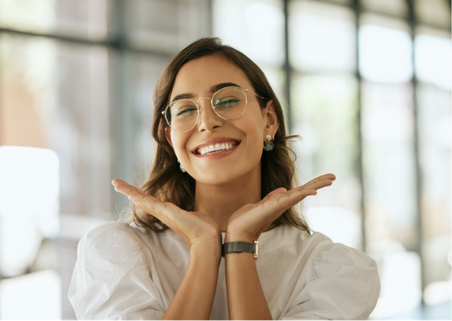 A smiling woman with glasses and earrings, posing with hands under her chin in a bright, modern office environment.