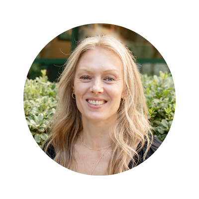 A woman with long blonde hair smiling outdoors, wearing gold hoop earrings and a black top, with greenery in the background who provides OCD treatment and therapy in San Diego, CA. 