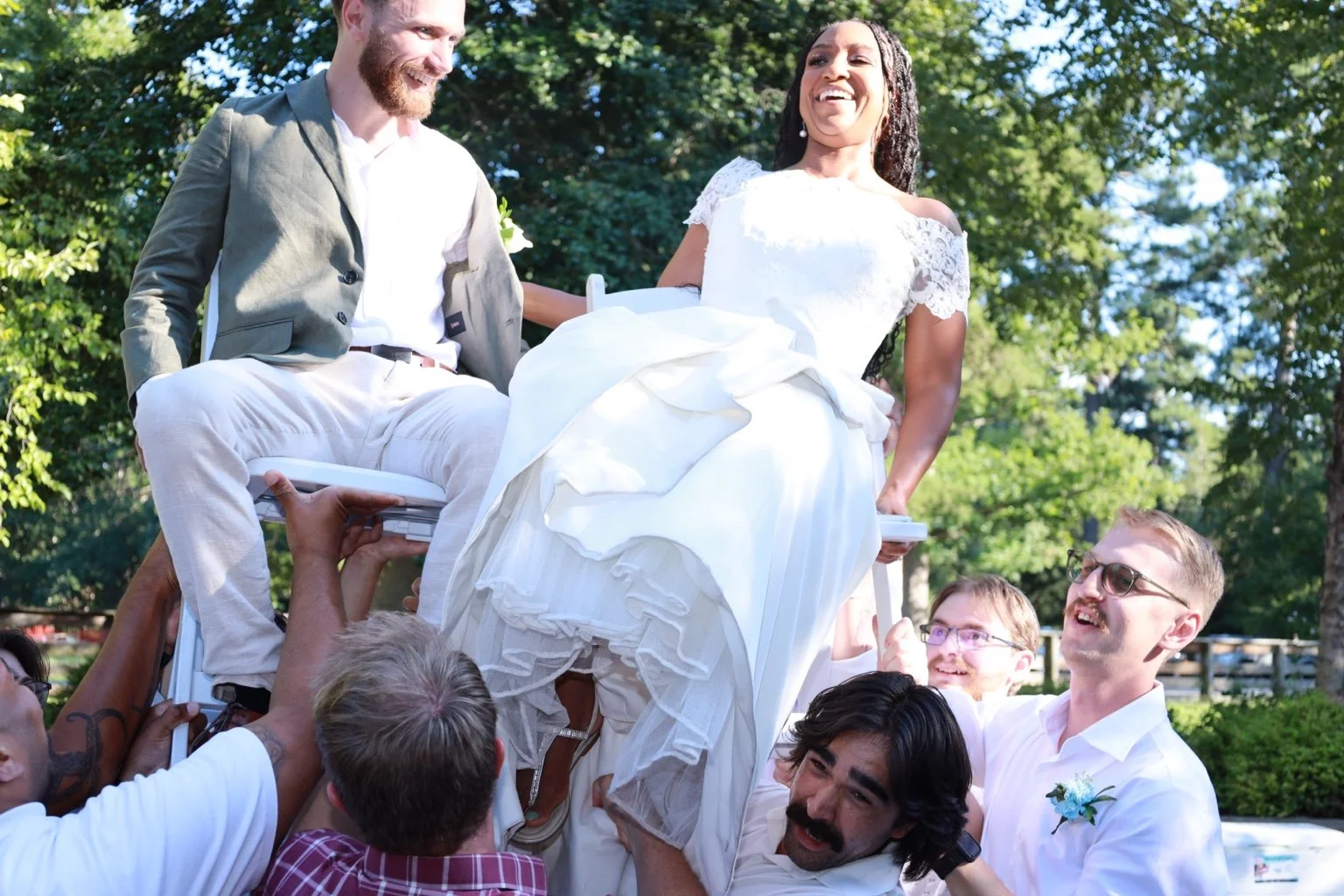 The bride and groom are lifted in celebration by their wedding party outdoors.