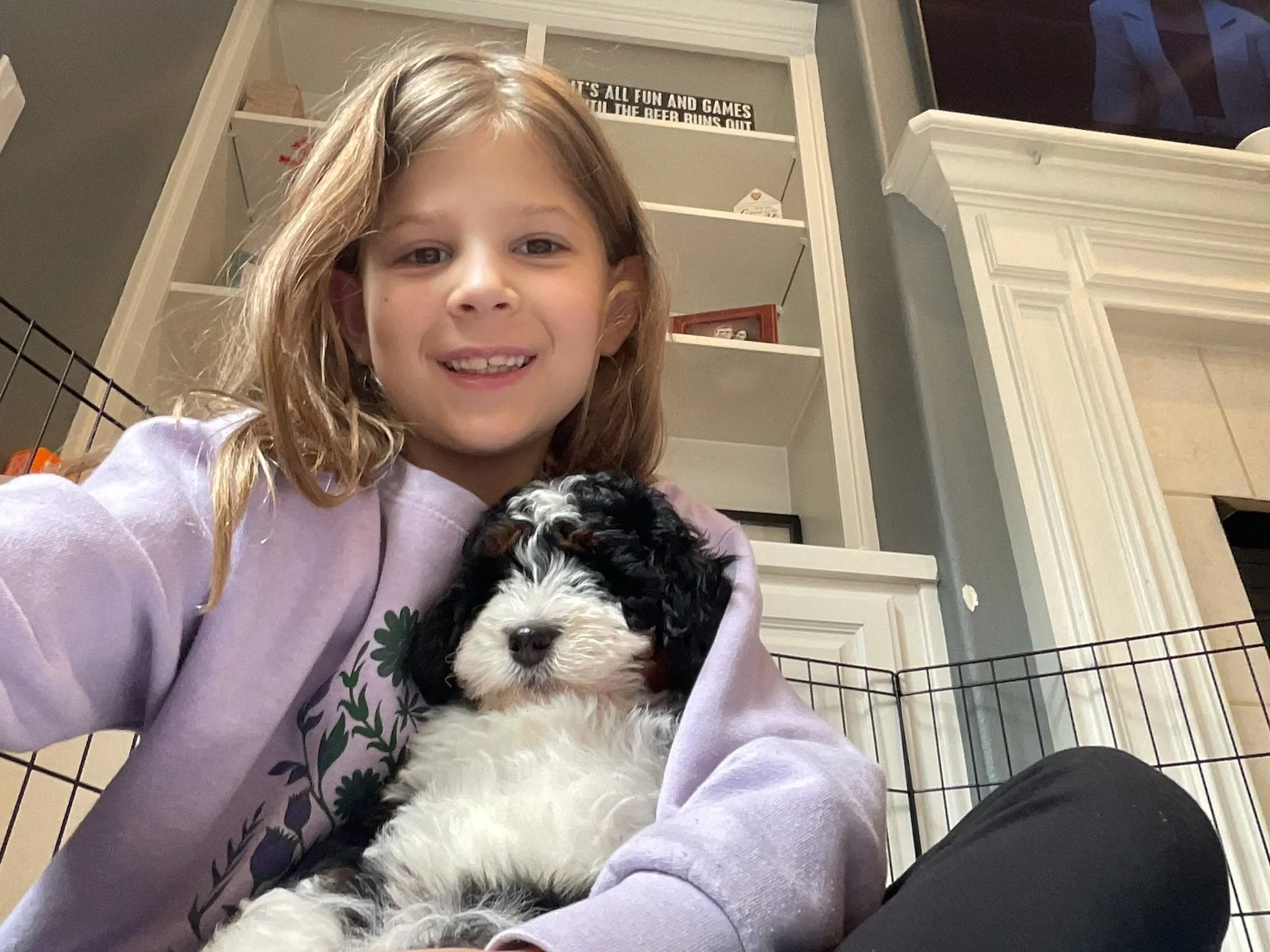 A young girl with light brown hair smiling while holding a black and white puppy, with a metal pet crate and white mantle in the background.