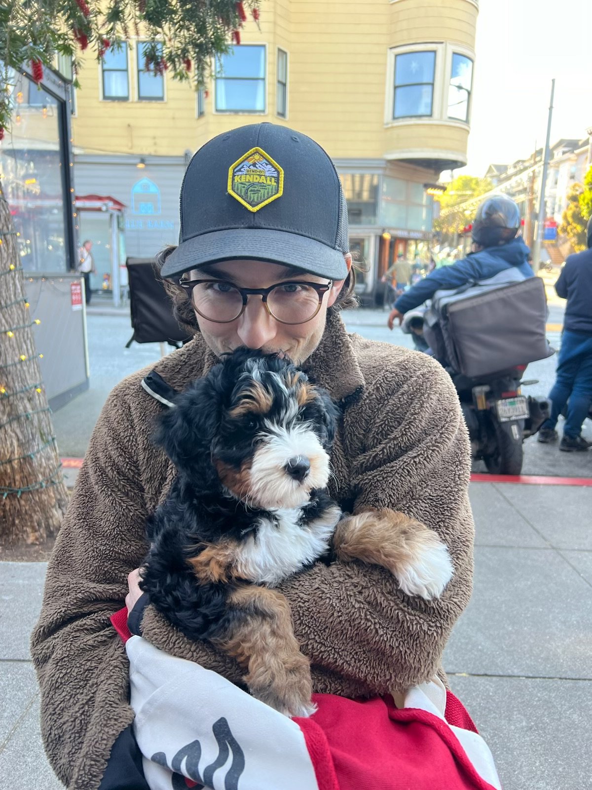 A young man with glasses and a brown fleece jacket holding a small black, white, and brown puppy, outdoors in an urban setting with buildings, people, and a motorcycle in the background.