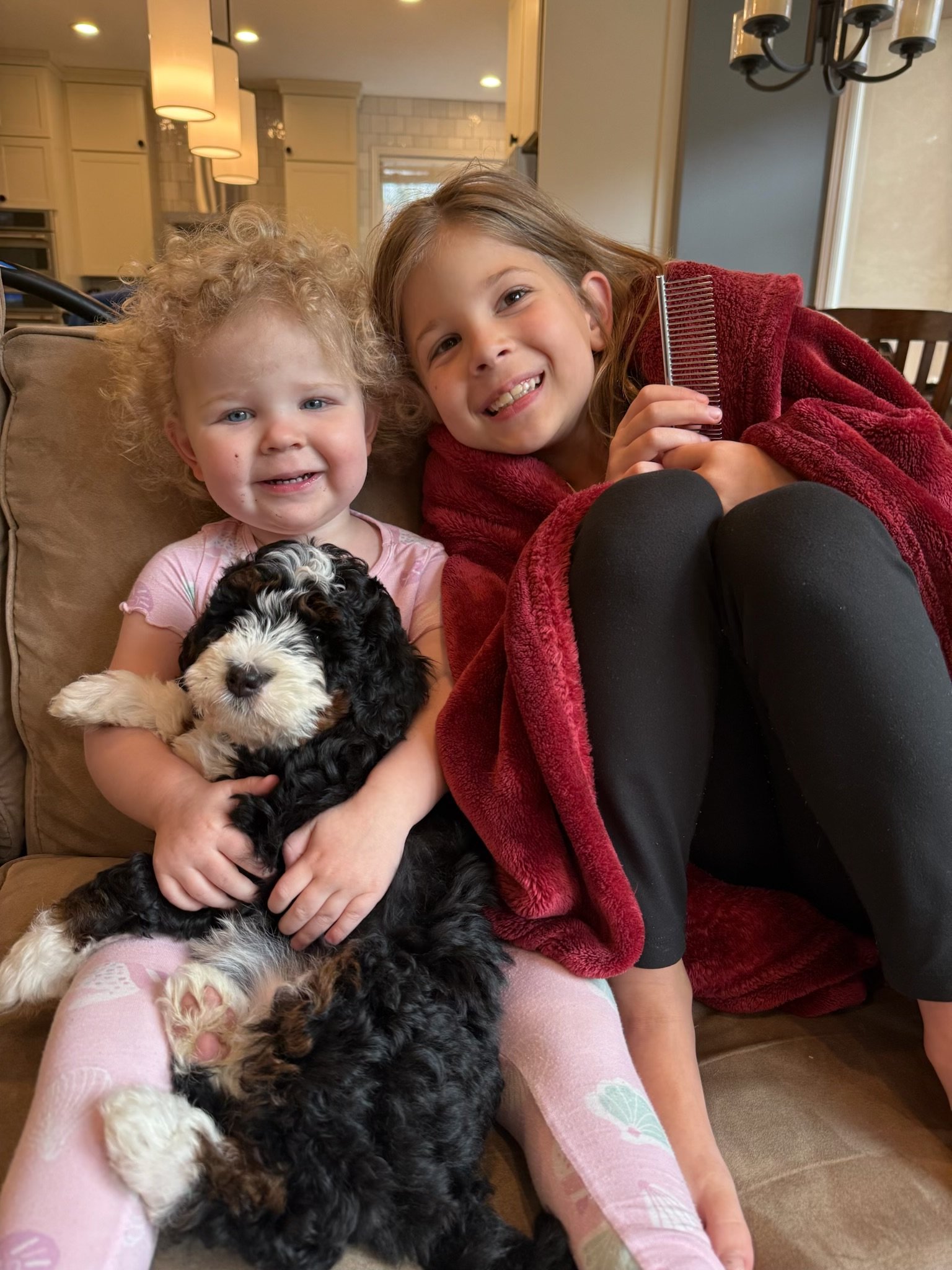 Two young girls sitting on a couch, smiling, with a black and white puppy on their lap, in a cozy kitchen setting.