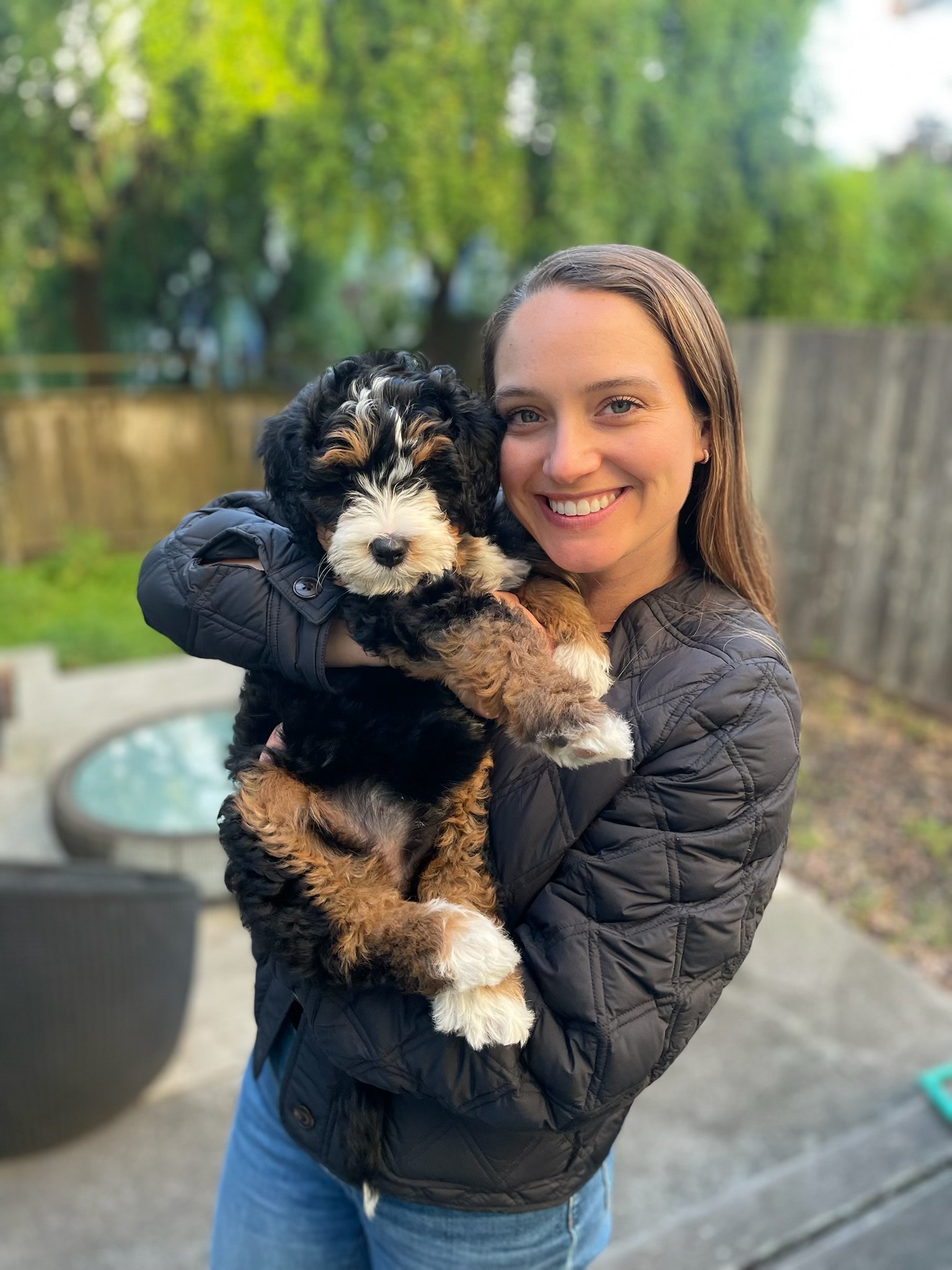 A woman with long brown hair and a black quilted jacket smiling while holding a fluffy black and white puppy outdoors with green trees and a wooden fence in the background.