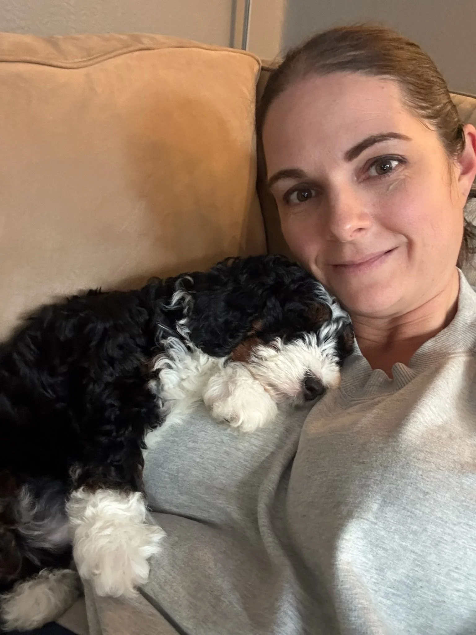A woman lying on a couch with a black and white curly-haired puppy resting her head on her face.