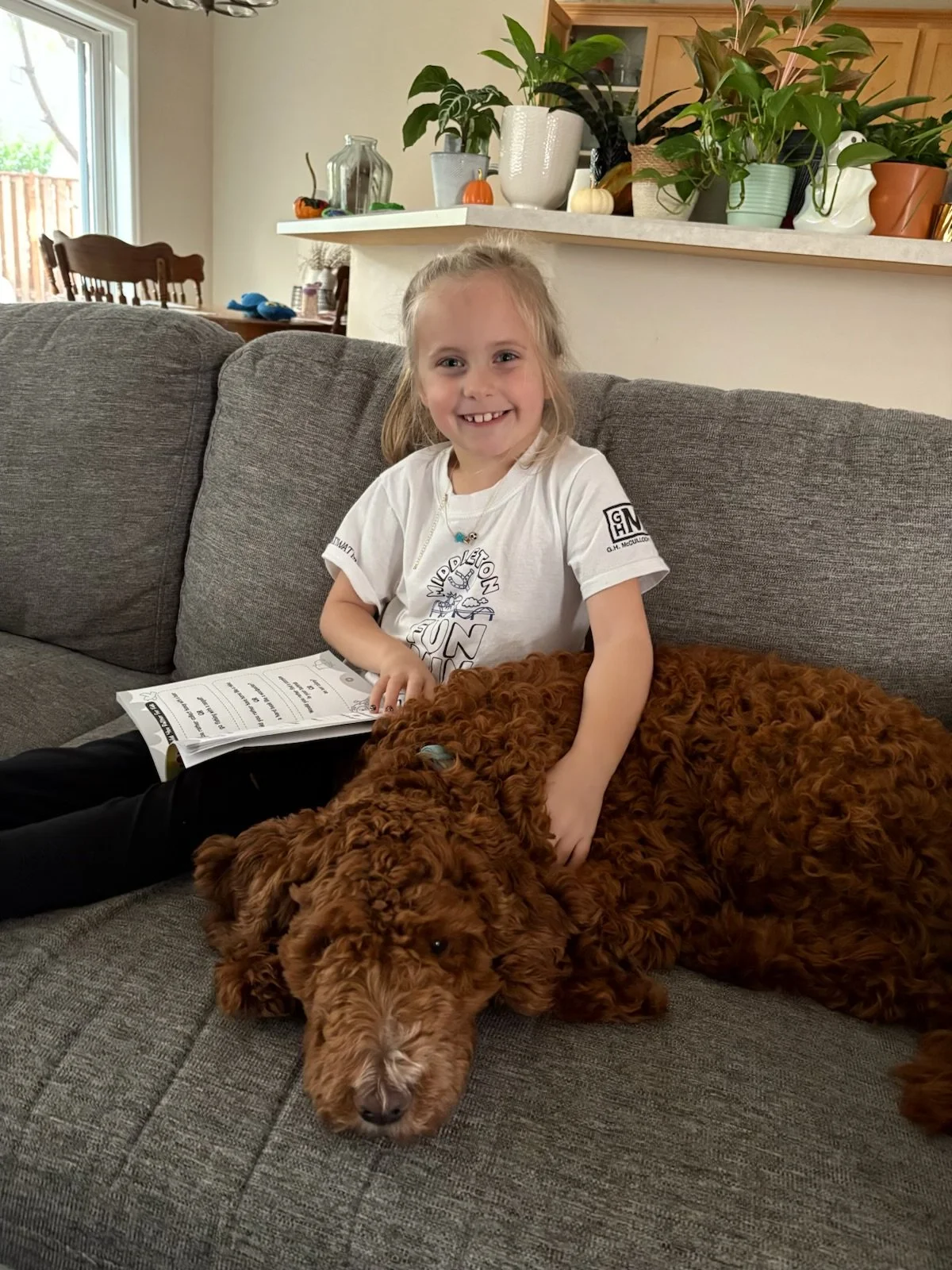 A young girl sitting on a grey couch with a large, curly-haired brown dog lying beside her. She is smiling and holding a book, indoors in a living room with plants and kitchen visible in the background.