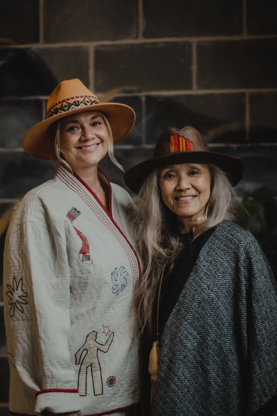 Two women wearing hats, one in a light-colored embroidered jacket and the other in a dark cloak, smiling in front of a dark brick wall.