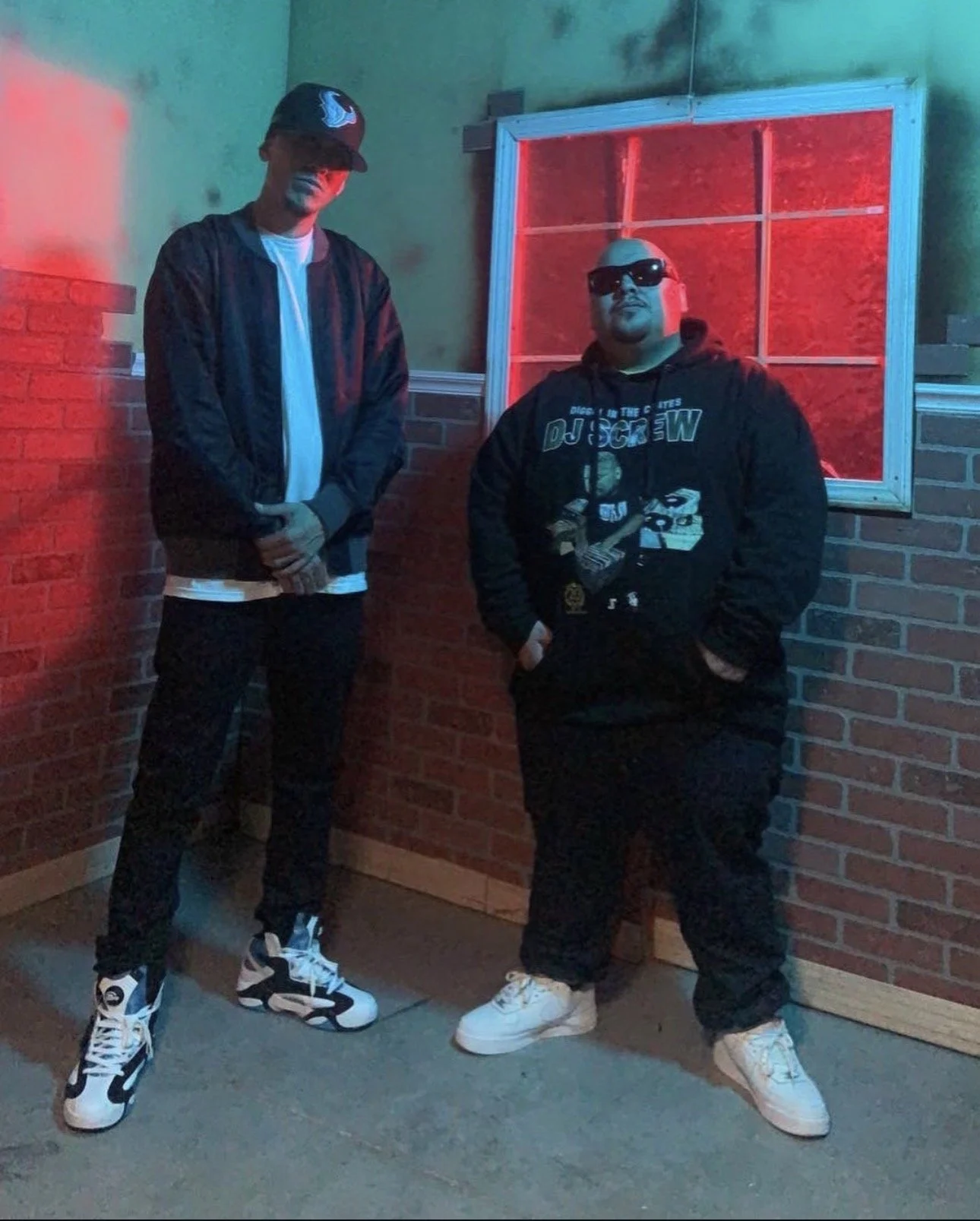 Two men standing against a brick wall at night, wearing streetwear and sunglasses.