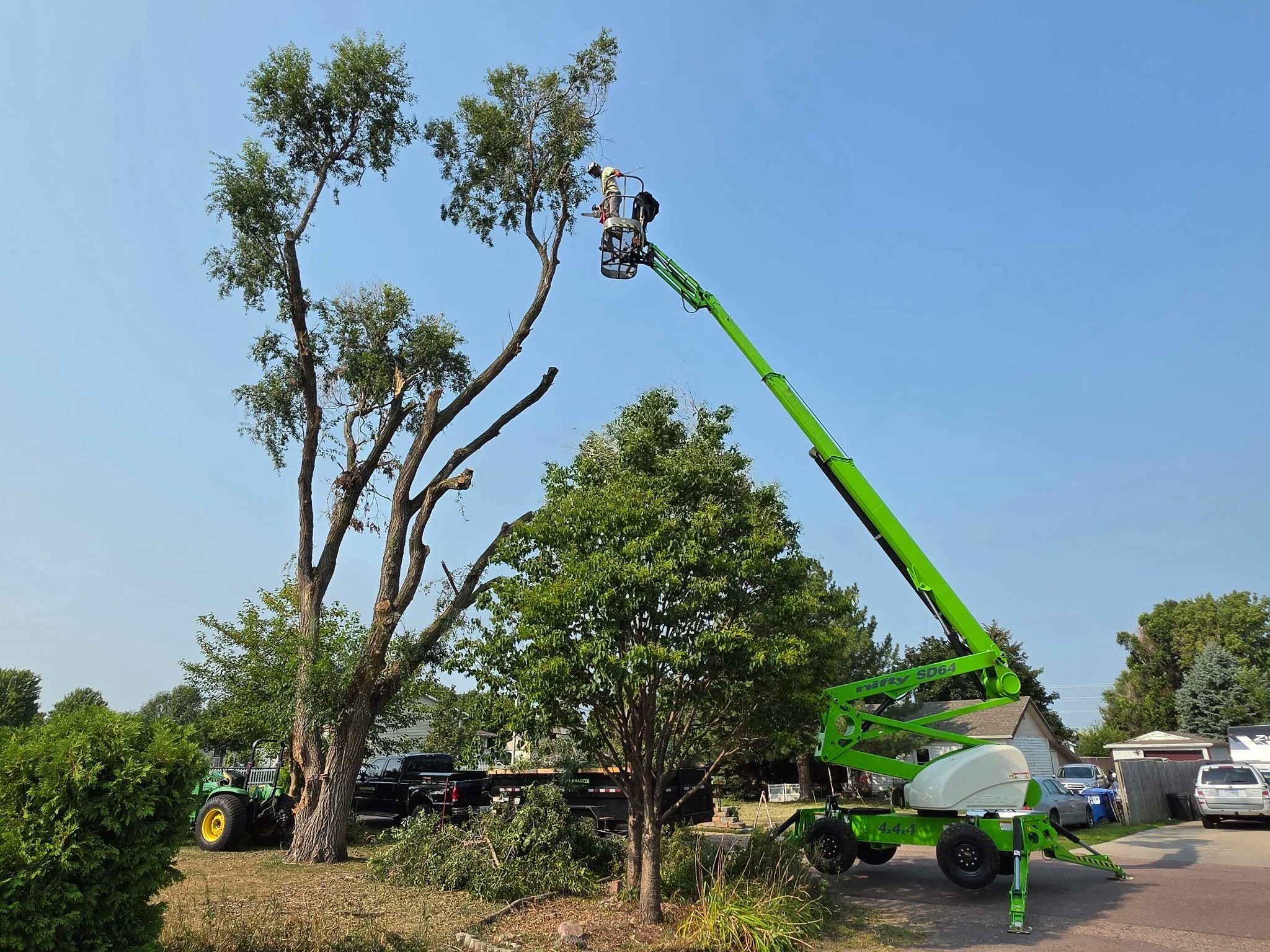 Tree trimming service using a green lift to trim branches in front of a house, with workers present and a service vehicle parked nearby.