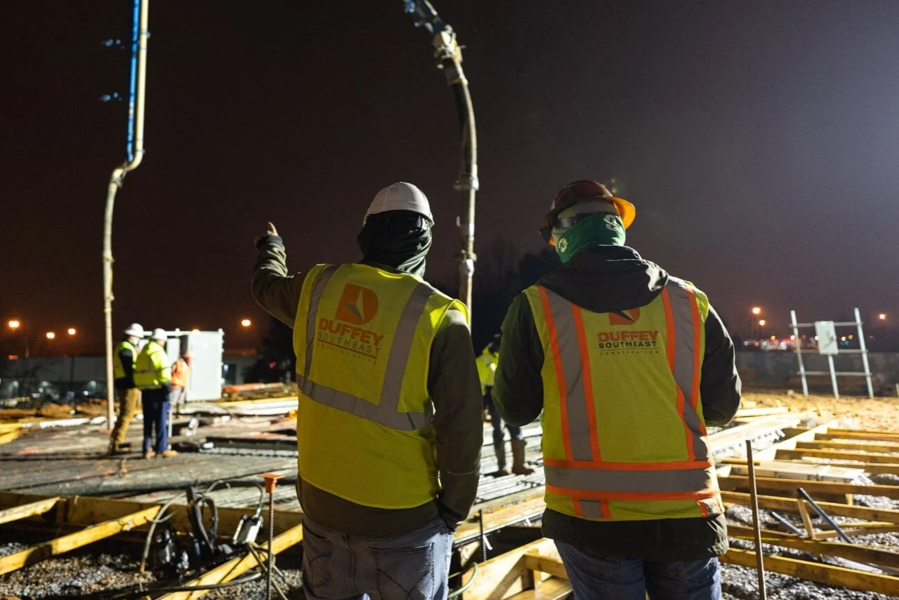 Construction workers at night on a building site, wearing safety vests, helmets, and high-visibility clothing, with some discussing or inspecting equipment.