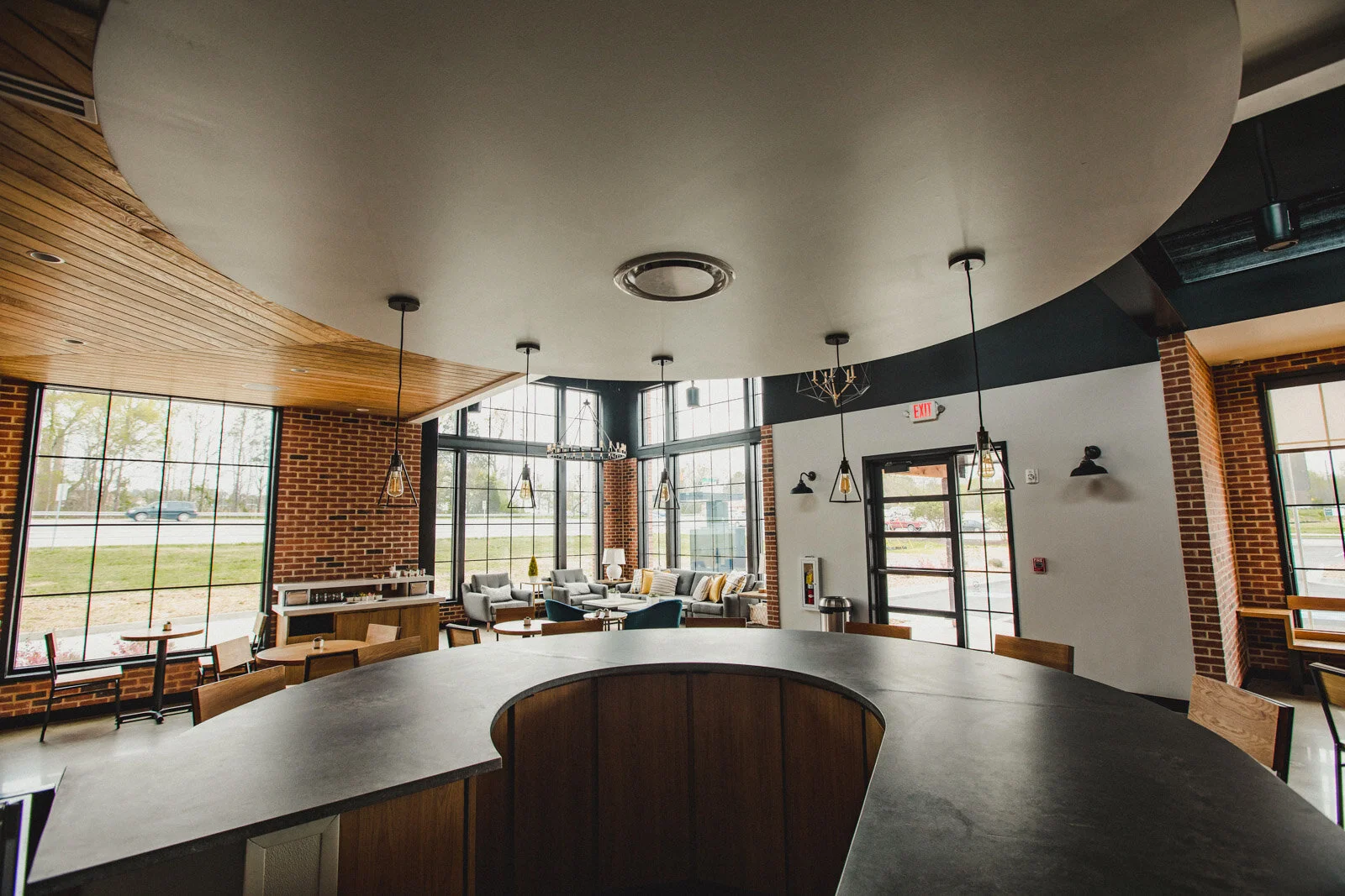 Interior of a modern cafe with large windows, brick walls, wooden ceiling, hanging pendant lights, and a seating area with gray sofas and chairs.