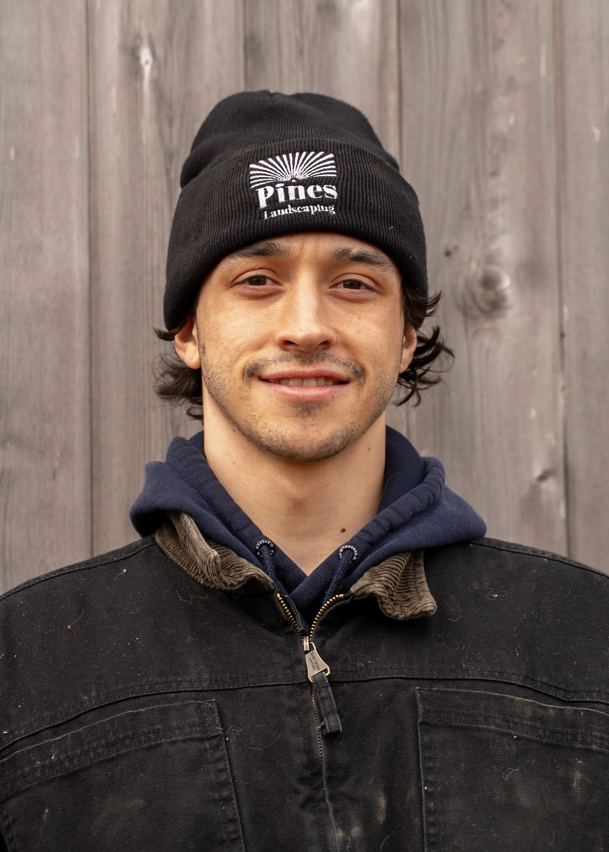 A young man with short dark hair, wearing a sleeveless black shirt with a 'Pines' logo, standing in front of a wooden fence.