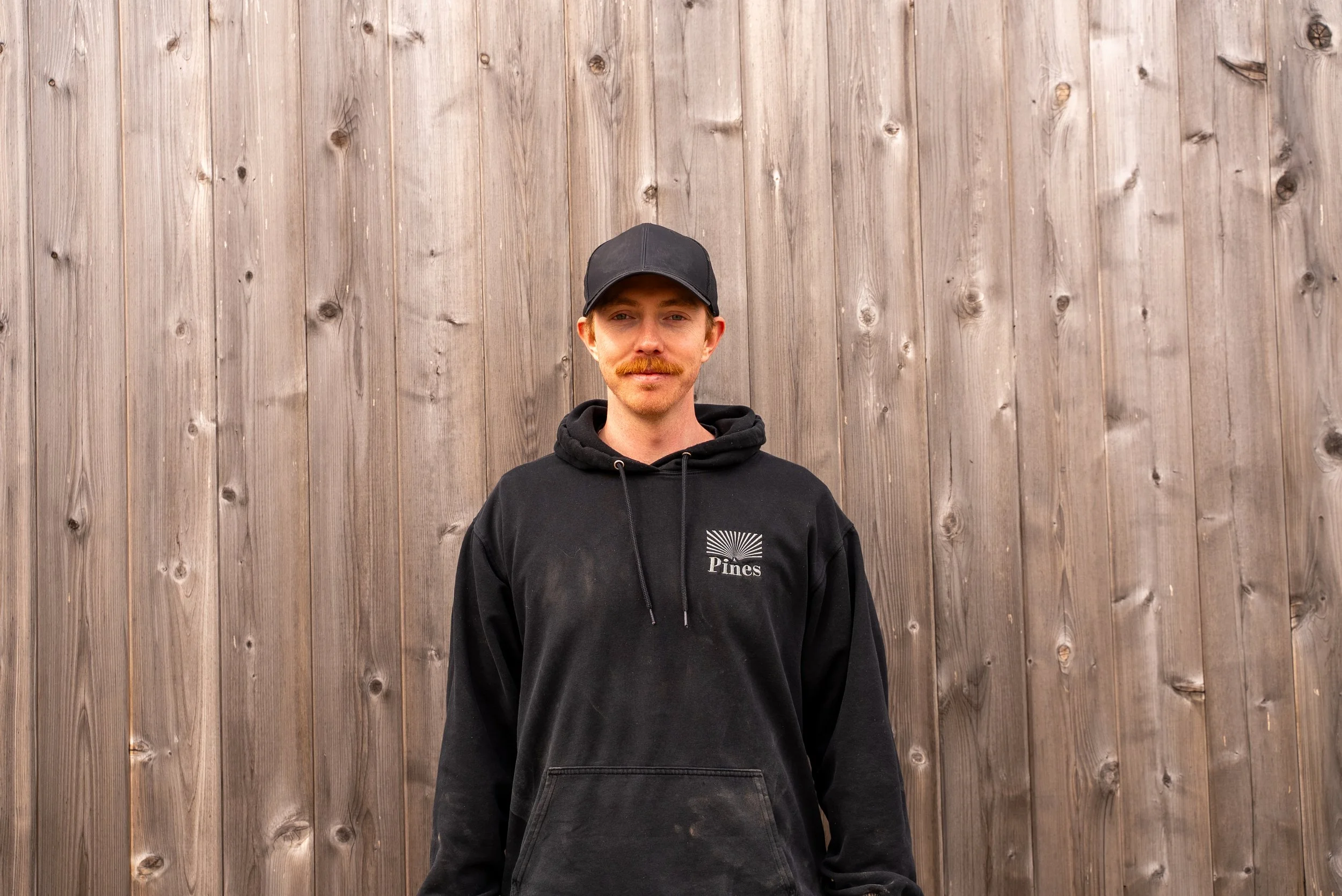 A young man smiling stands in front of a wooden fence, wearing a tan baseball cap and a black hoodie with the 'Pines Landscaping' logo.