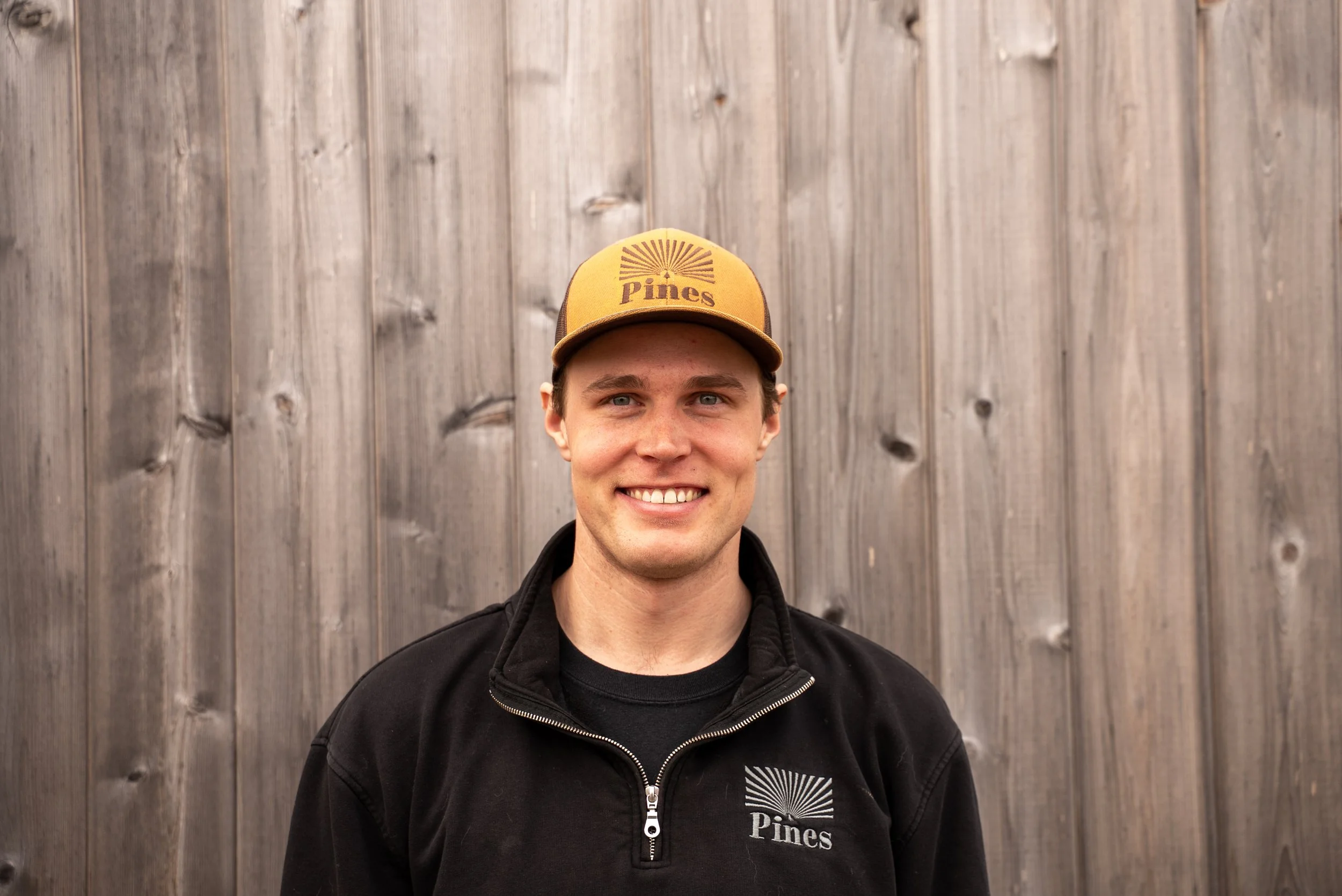 A young man standing in front of a wooden fence, wearing a black quarter-zip fleece with 'Pines' logo and a brown 'Pines' cap, smiling at the camera.