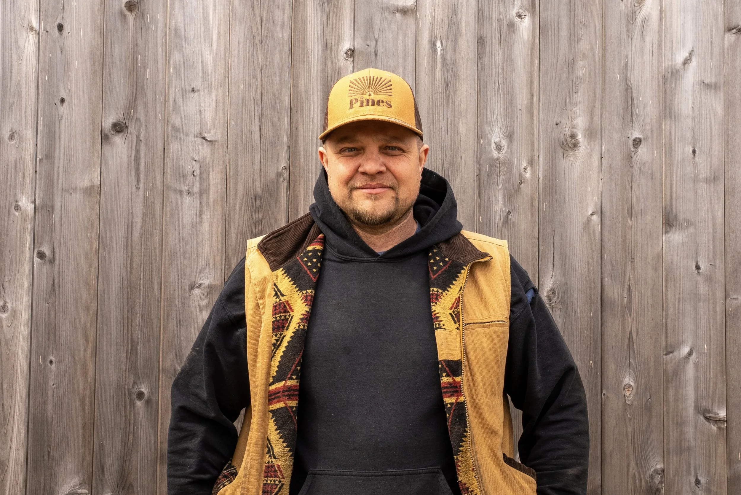 A young man wearing a tan cap and an olive green shirt with a logo reading "Pines Landscaping" standing in front of a wooden fence.