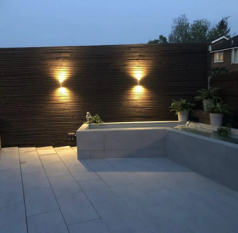 A modern outdoor patio area with a white tiled floor, potted plants on a ledge, and a dark wooden fence illuminated by two wall-mounted lights at dusk.
