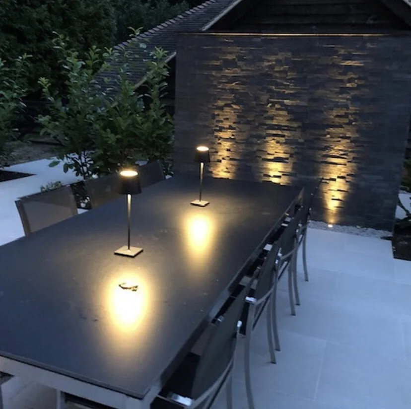 Outdoor dining table with six chairs and two small black table lamps, situated on a patio area with stacked stone wall and surrounding greenery, evening setting.