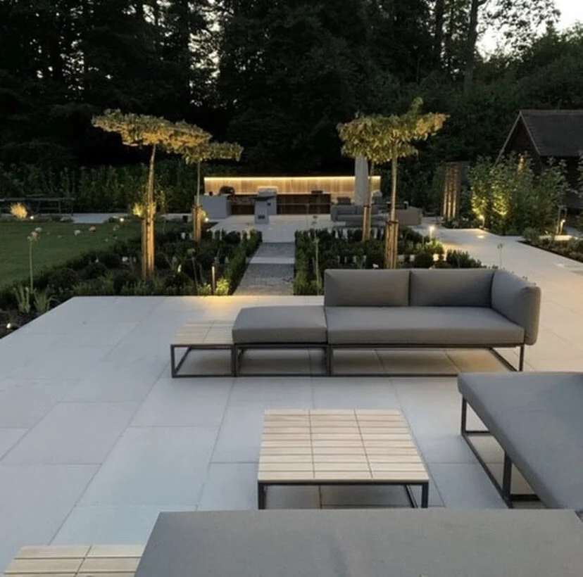 A modern outdoor patio at dusk featuring gray sectional sofas, small wooden tables, and evenly spaced young trees with canopy umbrellas. In the background, a landscaped garden with illuminated pathways and a sitting area  surrounded by dense trees.