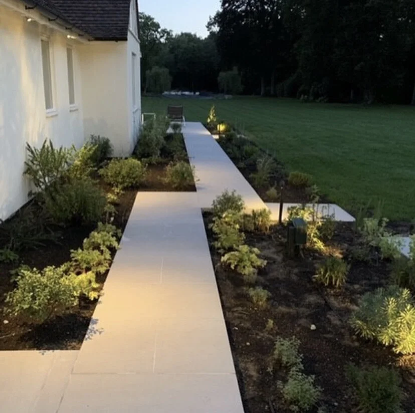 A well-lit backyard pathway with shrubs and plants along its sides, leading from the house down the garden to another patio area.