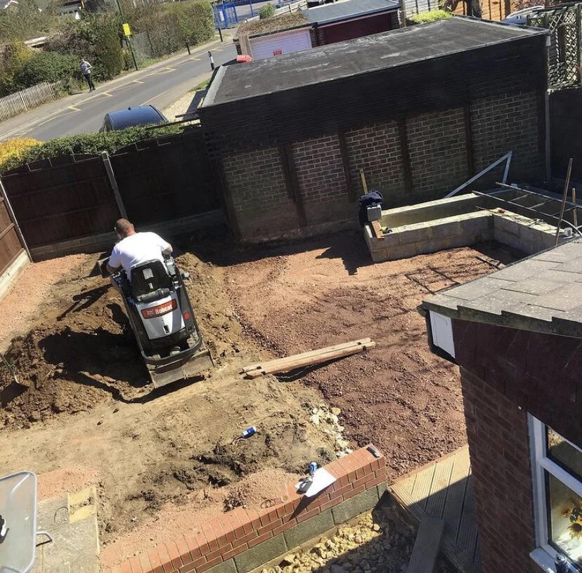 A person operating a mini excavator on a residential backyard construction site surrounded by brick walls and fencing, with a partially completed concrete structure in the background, and construction tools scattered around.