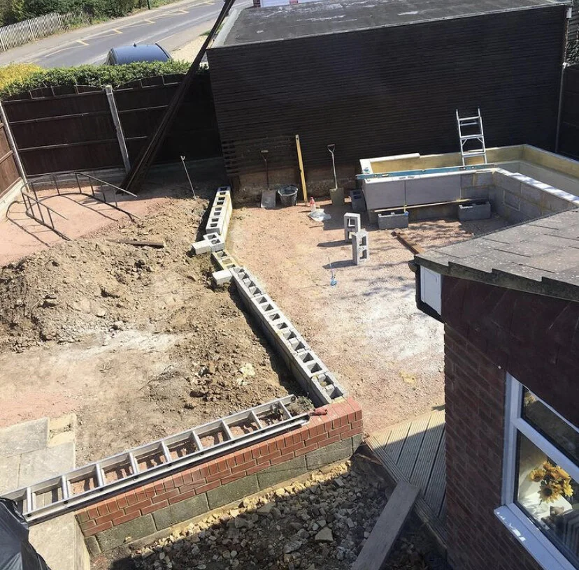 Backyard under construction with cinder blocks forming a raised garden bed, dirt area for future planting, a partially built brick wall, and a pool area with a ladder. Tools and construction materials are present.
