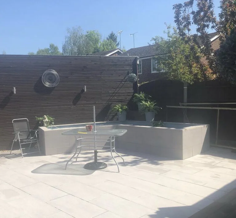 Patio with a glass table, metal chairs, a potted plant, and a raised concrete planter with plants. A wooden privacy fence and neighboring houses are in the background under a clear blue sky.